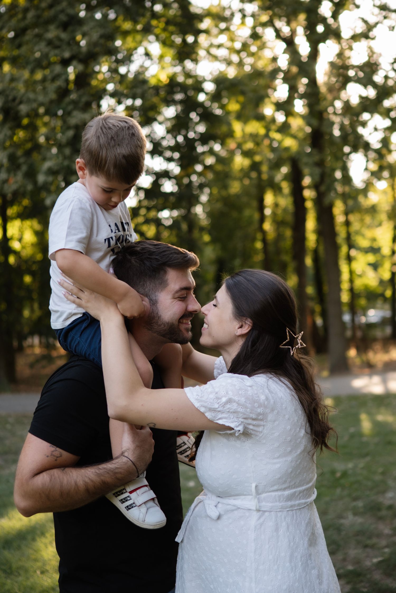 A happy family with a bearded dad carrying a young boy on his shoulders while a woman in a white dress embraces them in a park.
