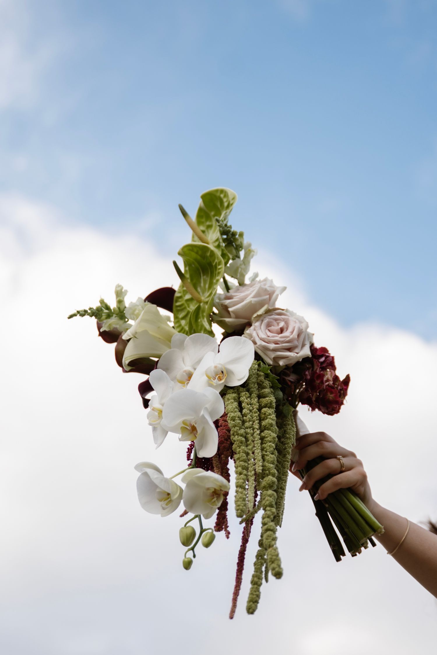 Hand holding a bridal bouquet with white orchids, roses, and greenery against a cloudy sky.