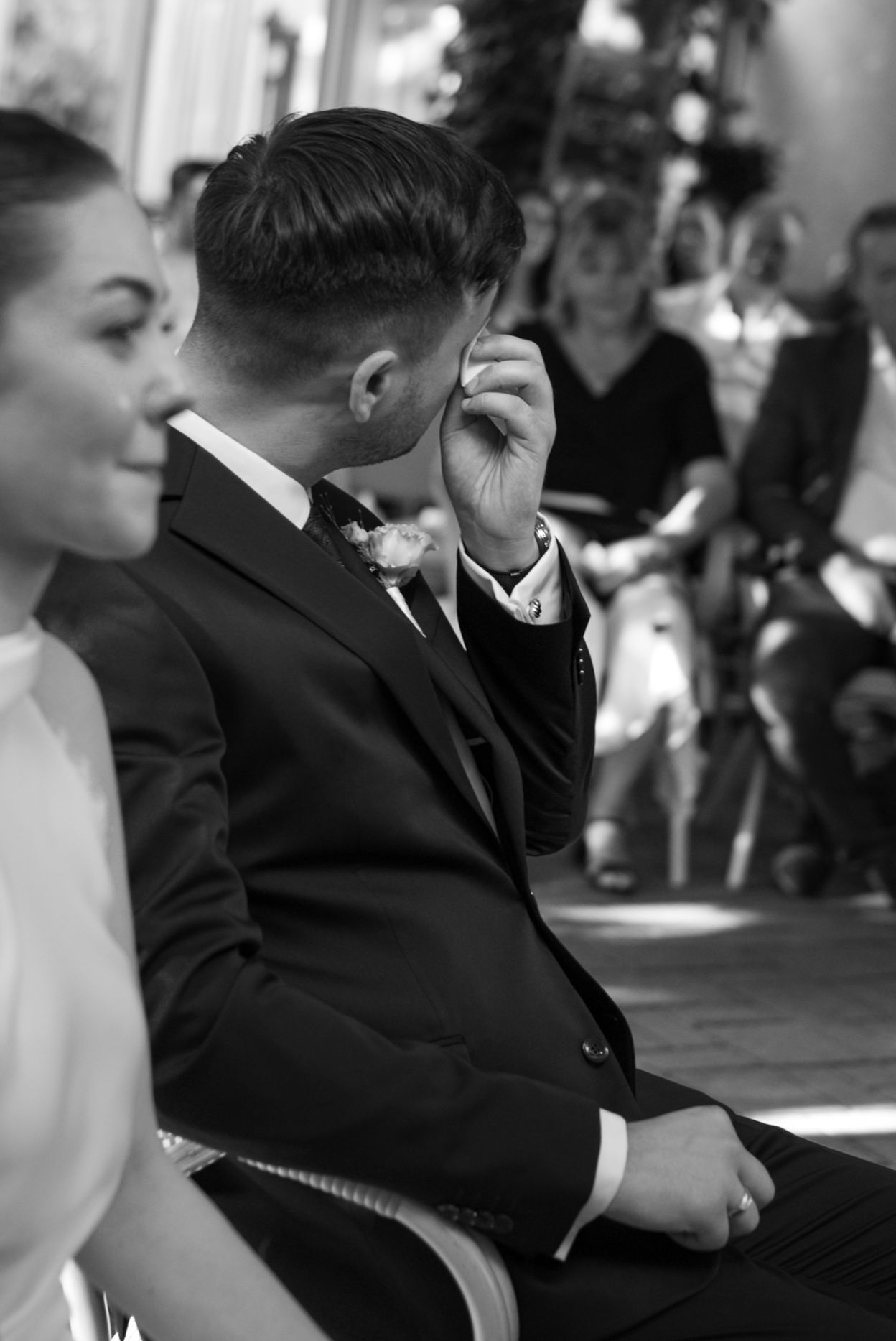Black and white photo of an emotional groom wiping tears during wedding ceremony while bride sits beside him.