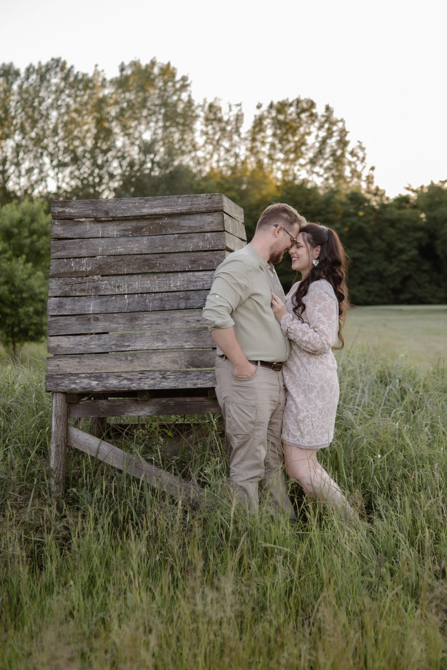 Couple sharing a romantic moment in a lush green field beside a rustic wooden structure at golden hour.