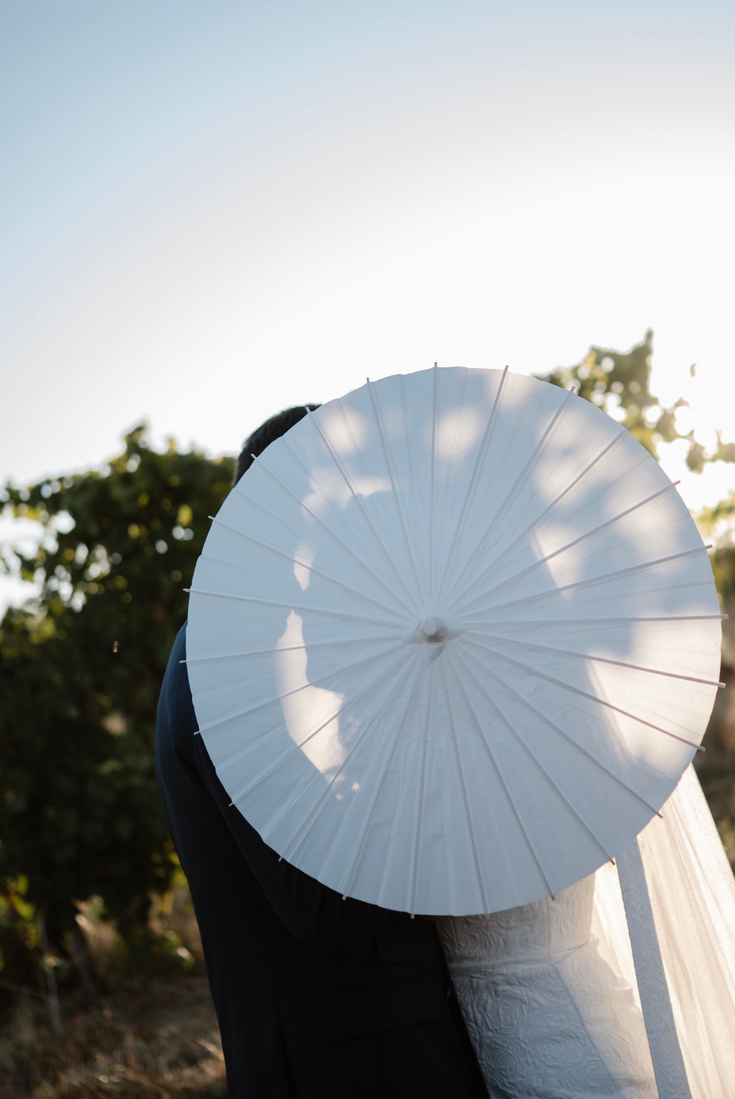 Couple sharing a kiss hidden behind a white parasol umbrella outdoors at a wedding ceremony.
