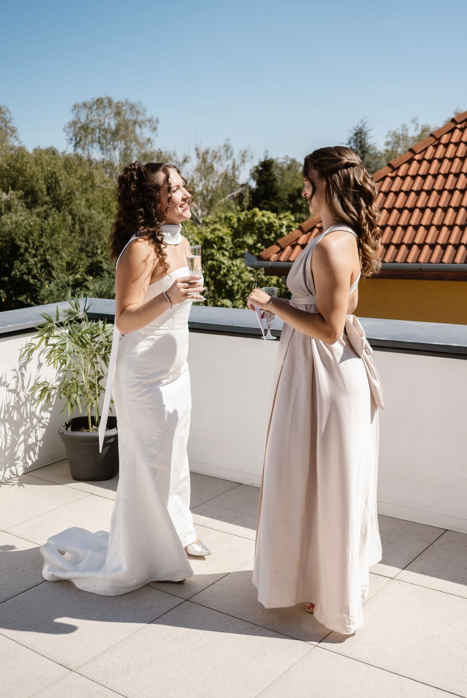 Bride and bridesmaid in elegant gowns toasting with champagne on a sunny rooftop terrace.