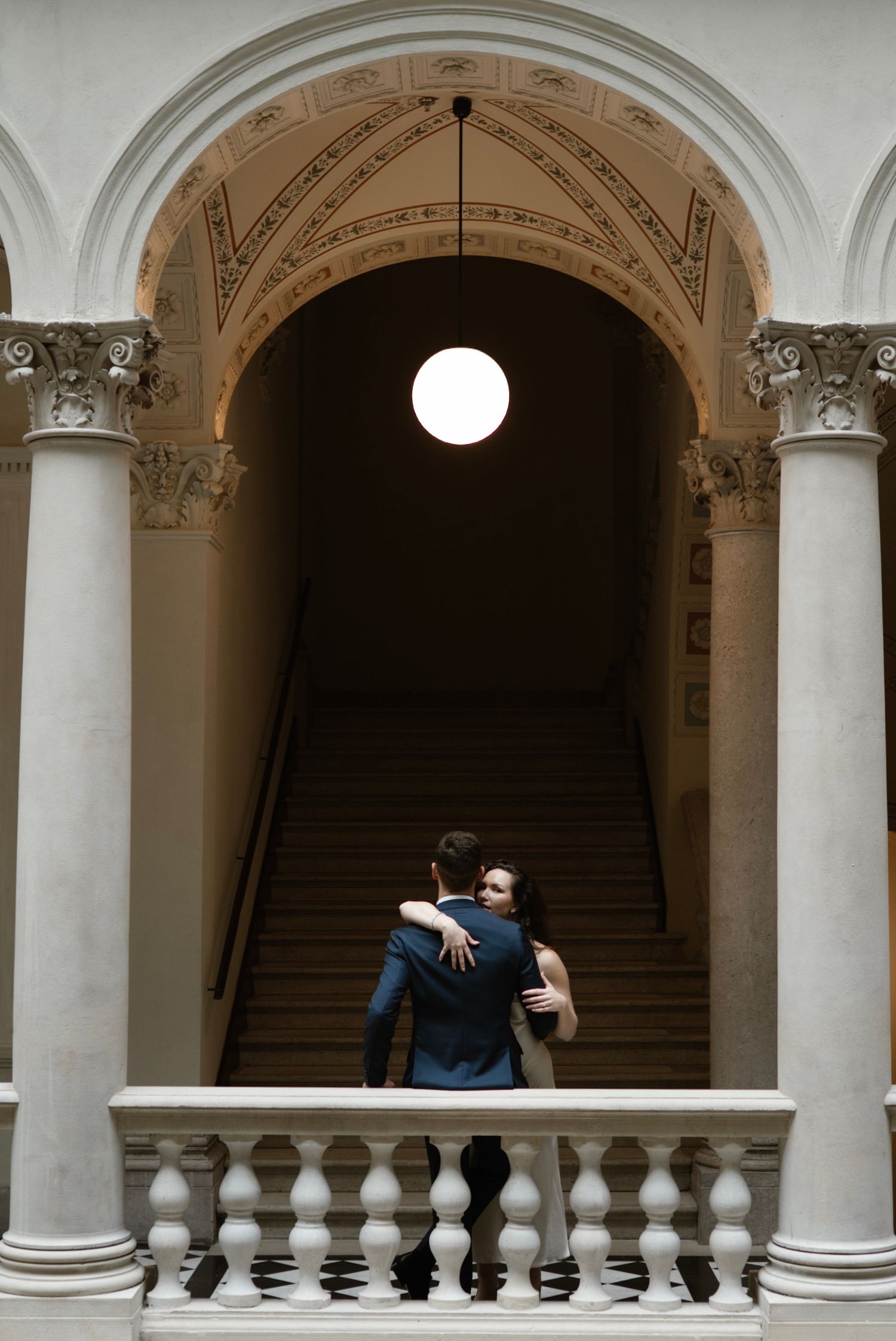 Couple embracing on elegant staircase under glowing pendant light, framed by ornate arch and white marble columns.