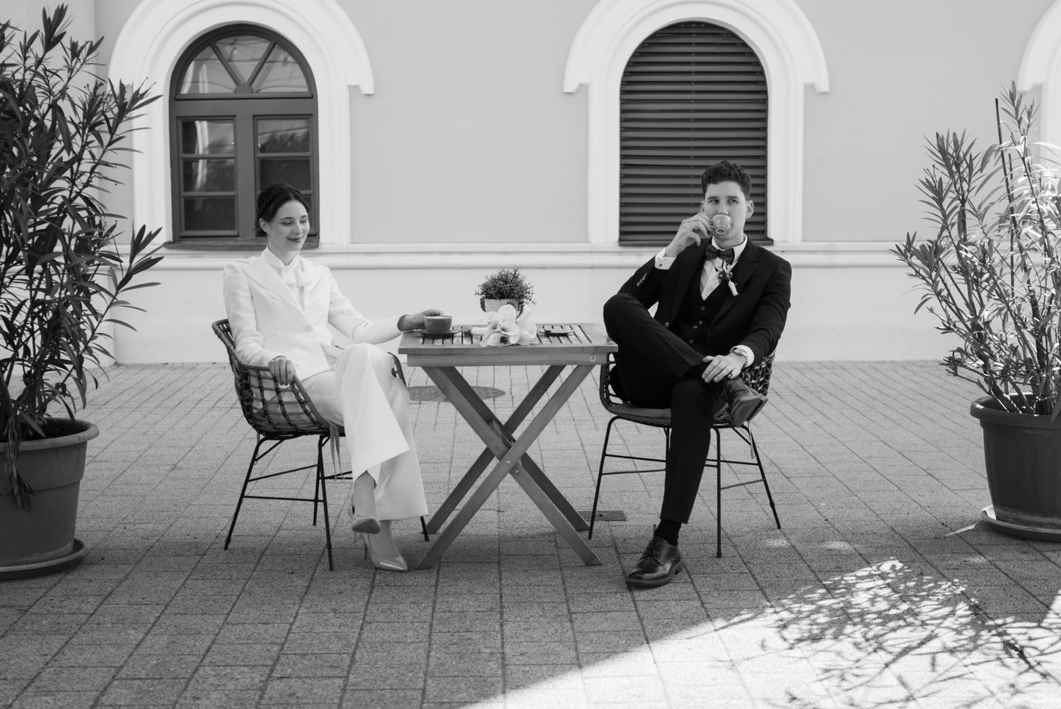 Black and white photo of a couple in formal attire sitting at a small outdoor table, posing for wedding portraits.