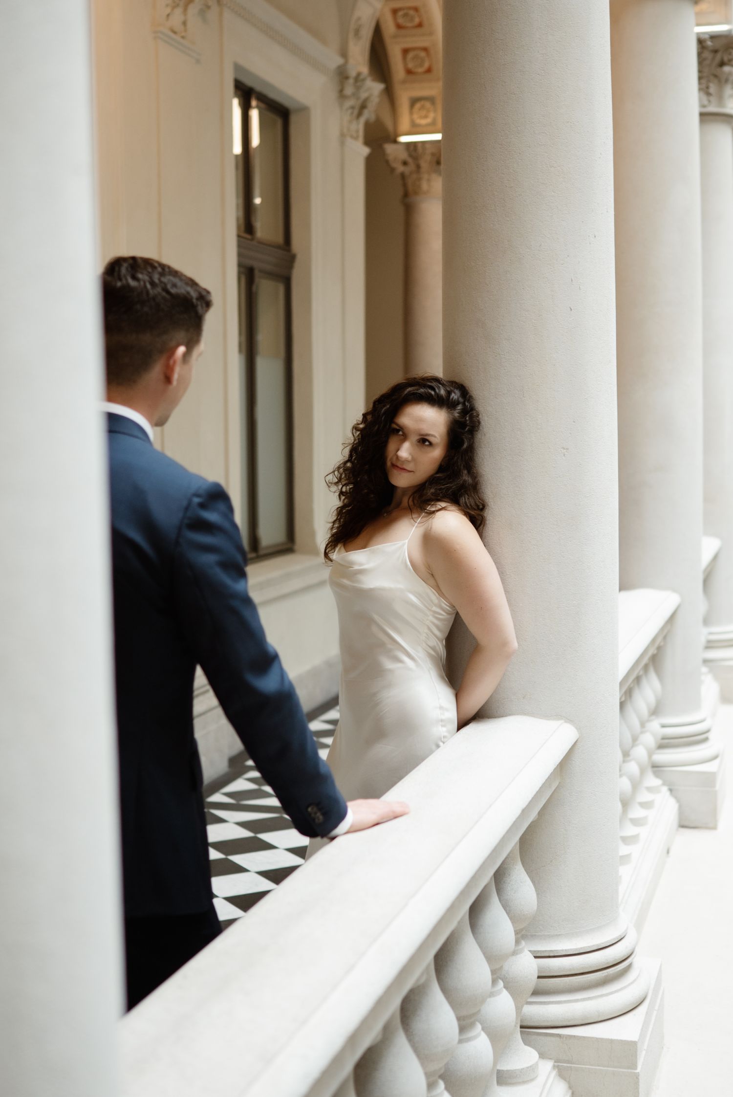Bride in white dress and groom in navy suit share a romantic moment on elegant marble balcony with columns.