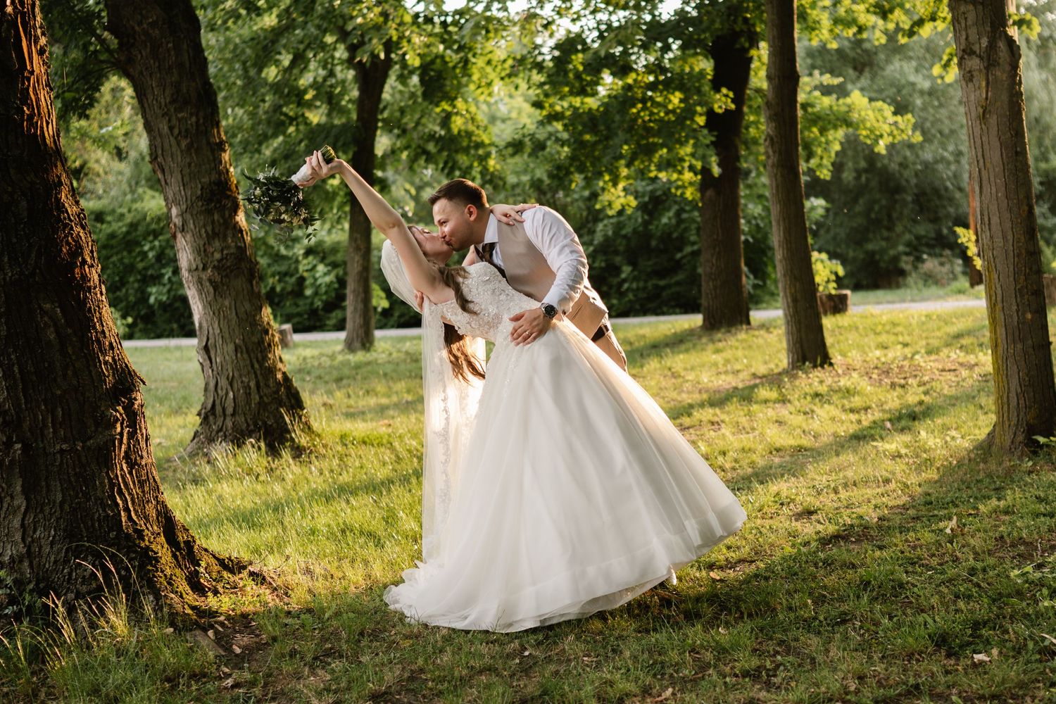 Wedding couple shares joyful moments under trees in a sunlit garden setting.