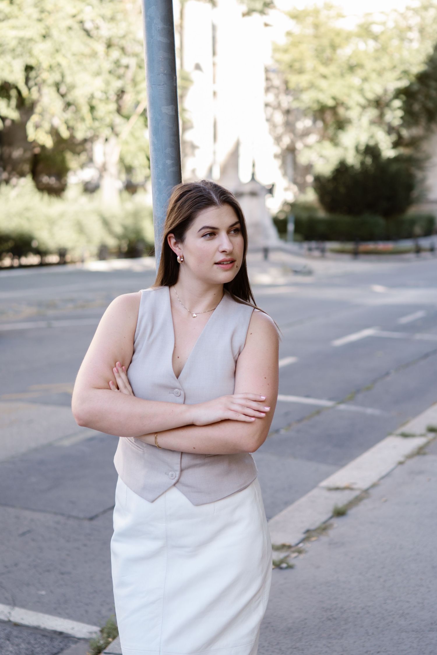 A person in a sleeveless gray and white top stands with crossed arms on a sunny street.