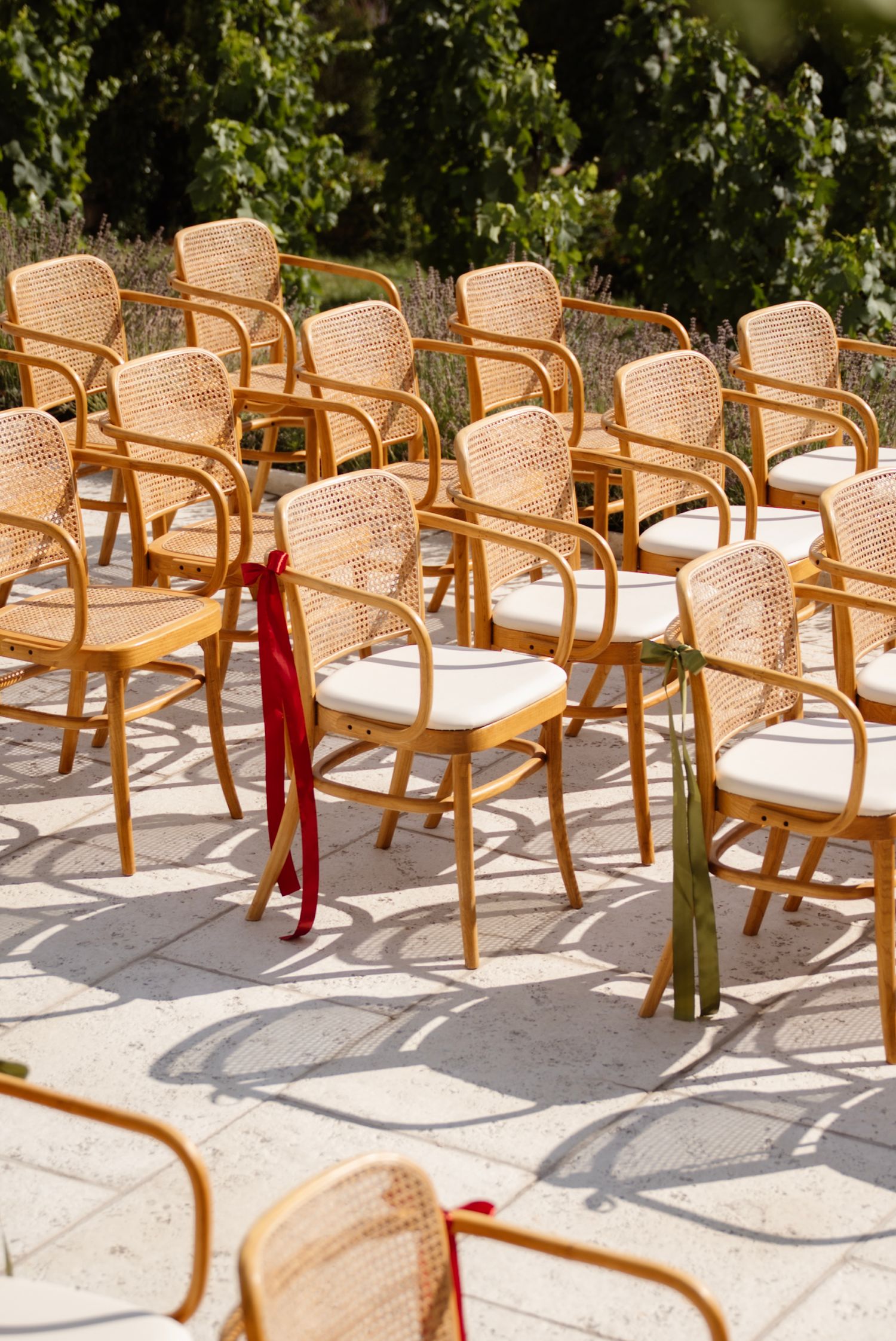 Rows of elegant rattan chairs with white cushions and ribbon accents set up outdoors for a wedding ceremony.