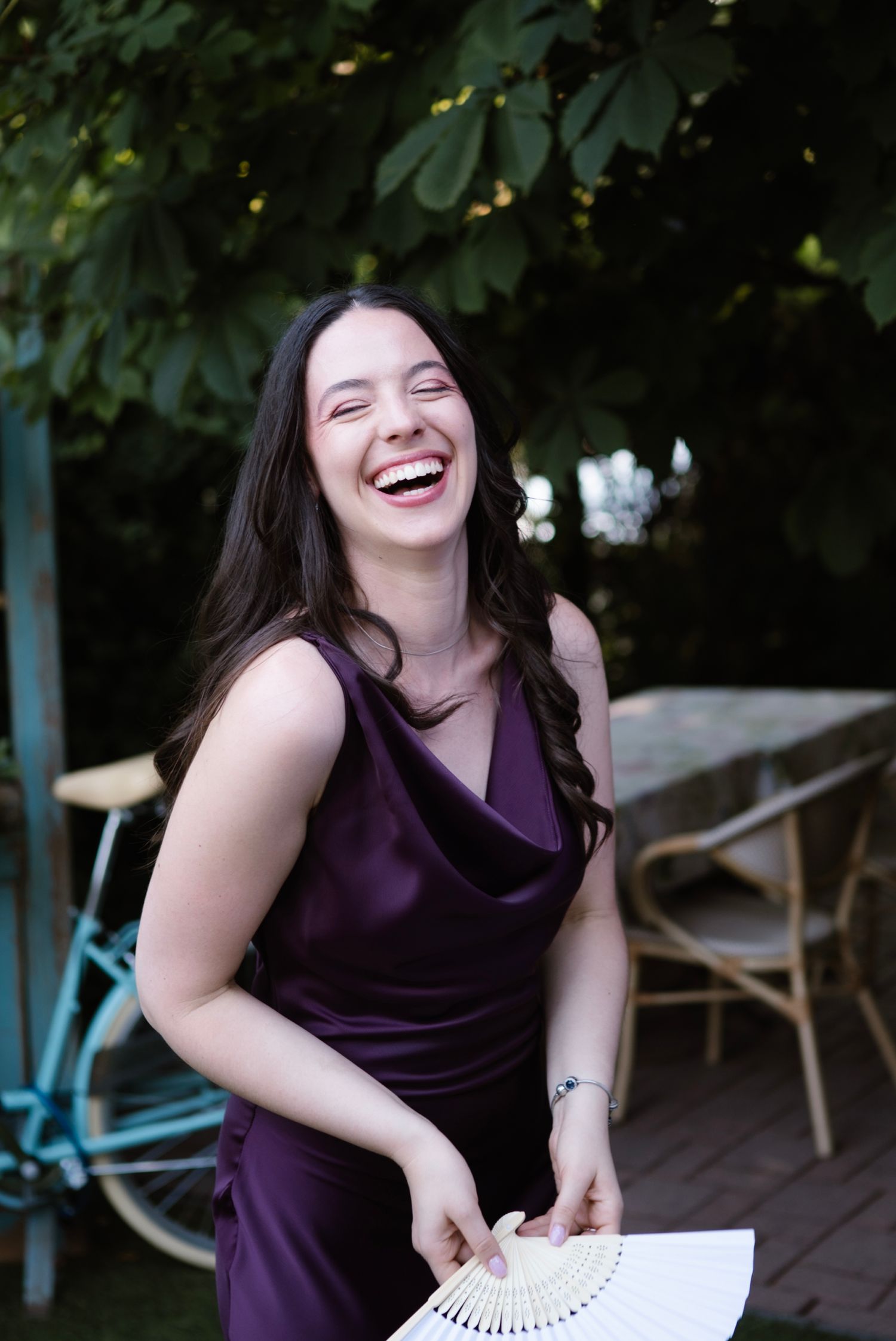 A laughing woman in a purple satin dress holds a white fan outdoors, with green trees in the background.