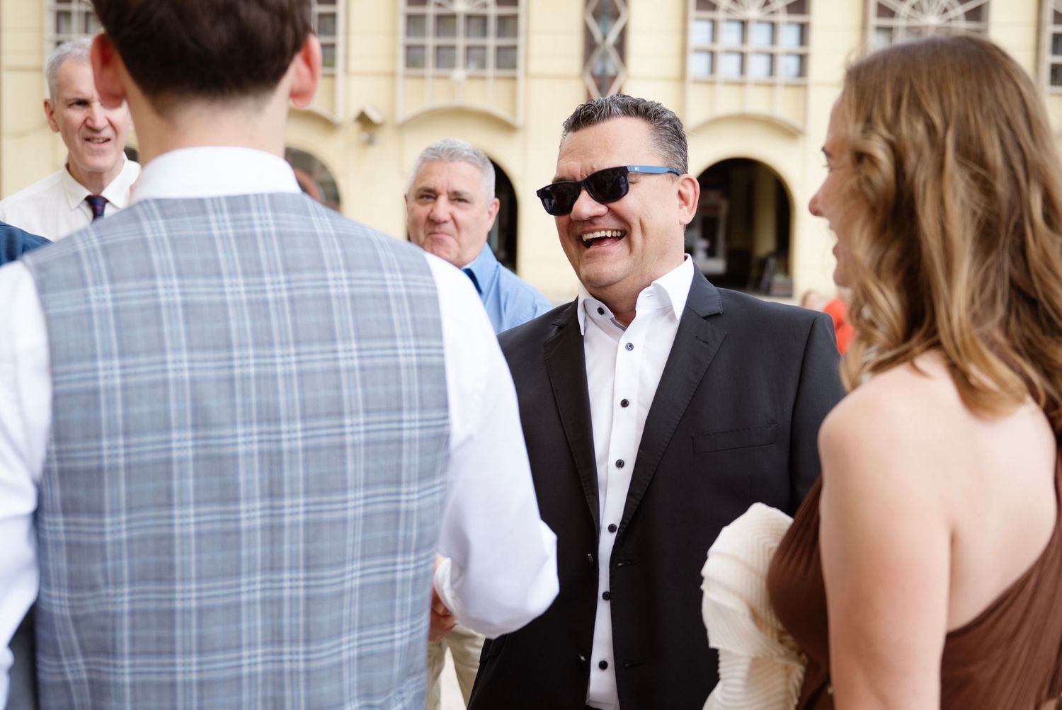 Man in black jacket and sunglasses laughing at outdoor wedding reception with guests in formal attire.
