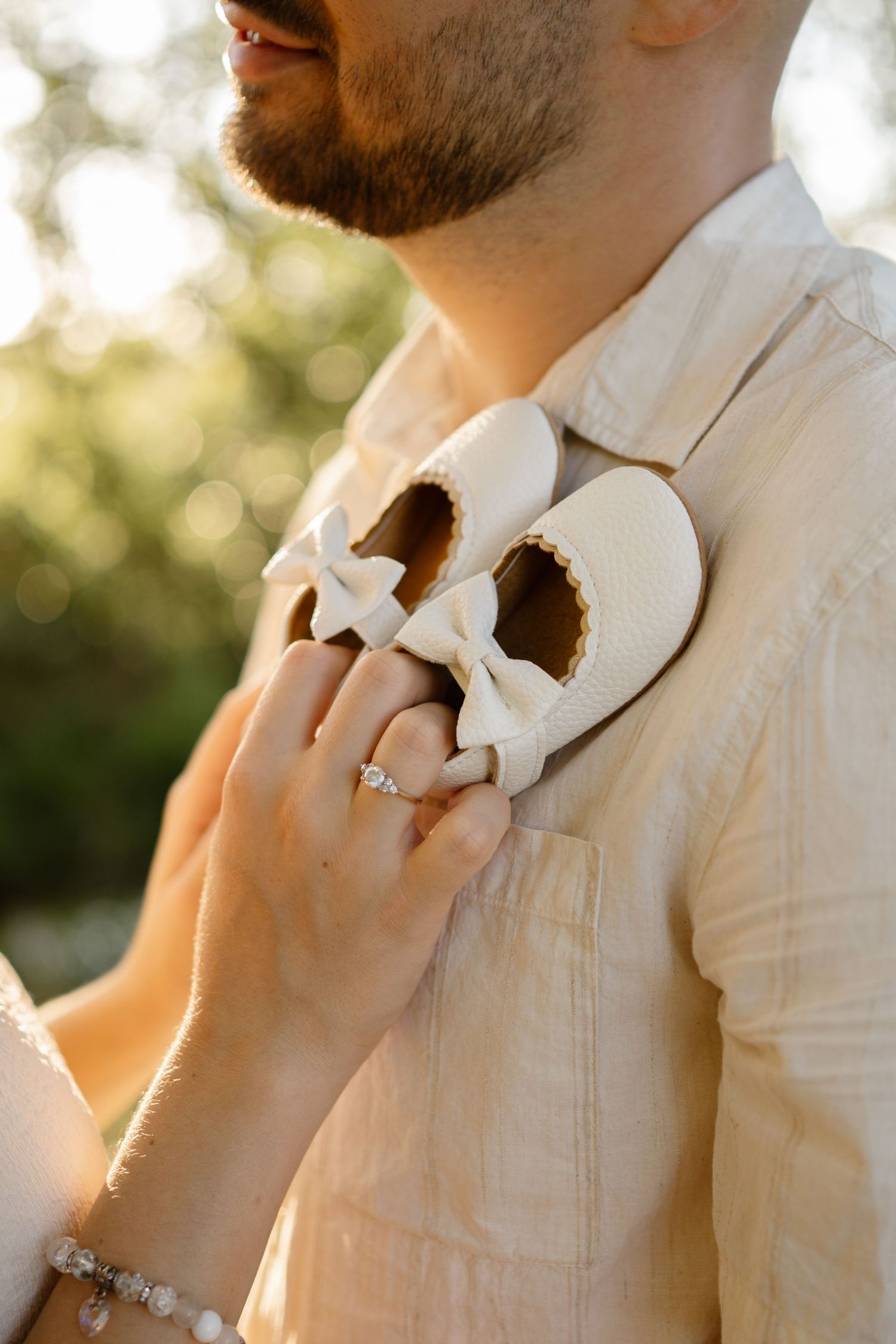 A close-up of tiny white baby shoes held against a white shirt in golden sunlight.