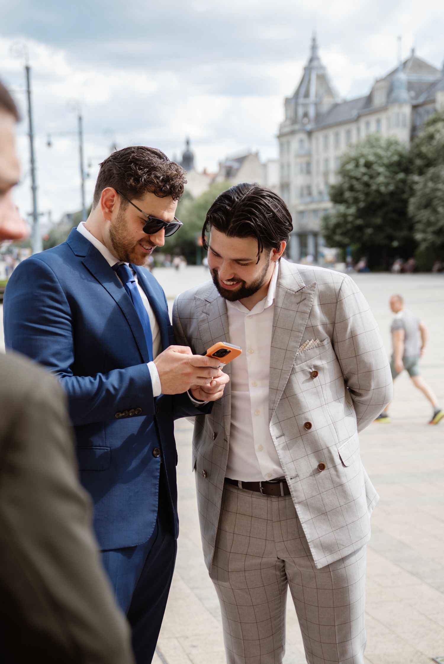 Two well-dressed businessmen in suits reviewing a smartphone outdoors in an urban plaza.