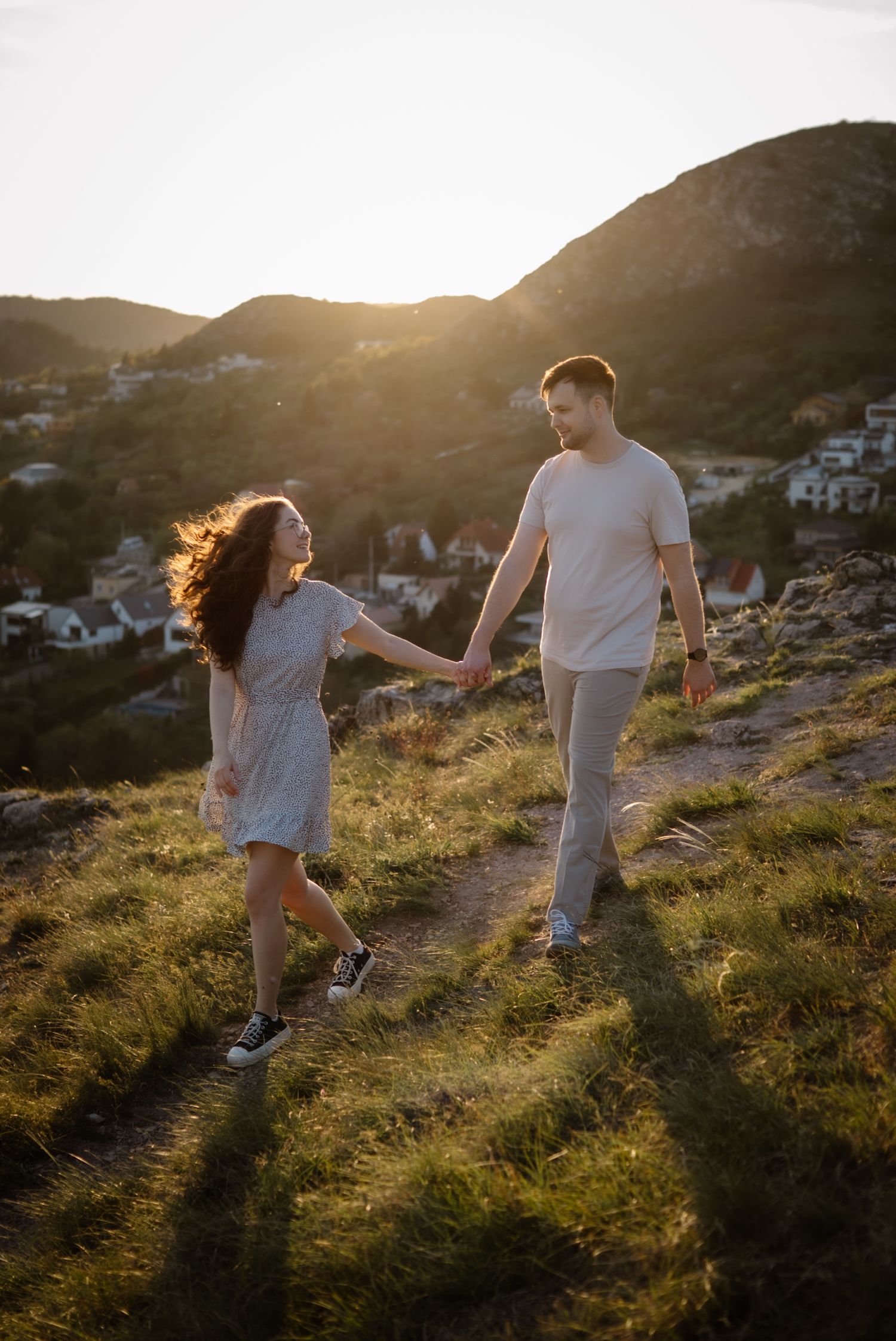 Couple holding hands walking on a grassy hillside at golden sunset with mountains in background.