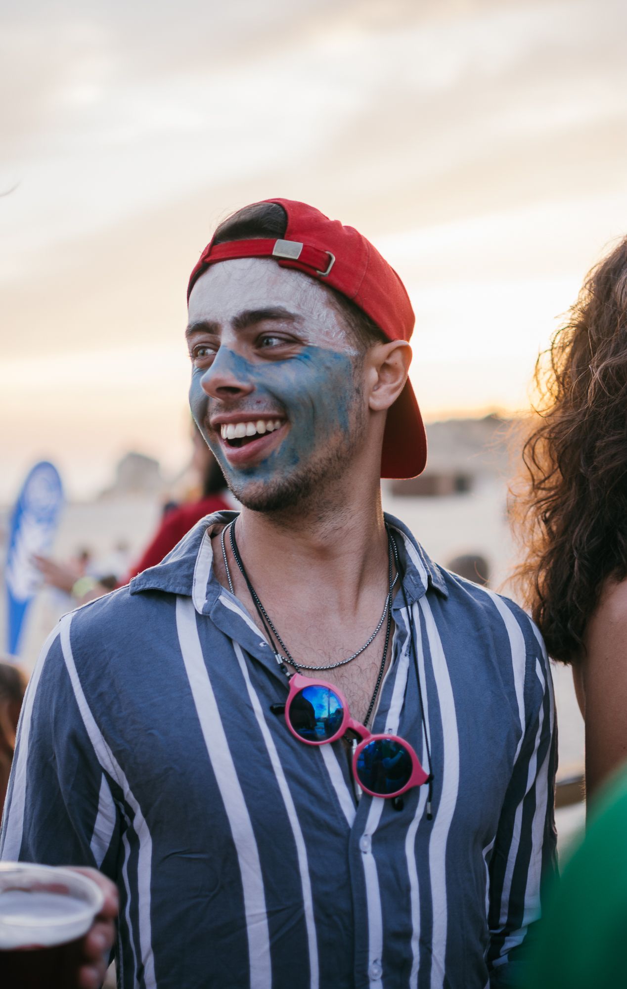 Someone wearing face paint and a red cap at an outdoor festival.