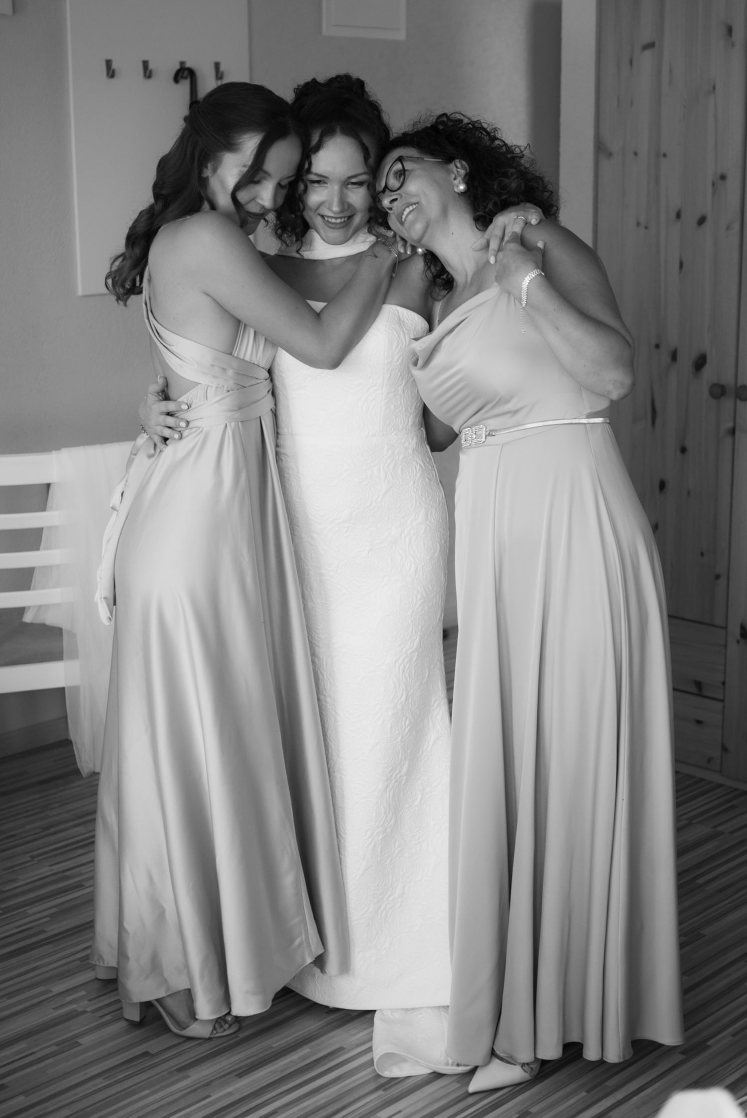 Bride in white gown embraced by two bridesmaids in elegant satin dresses, black and white photo.