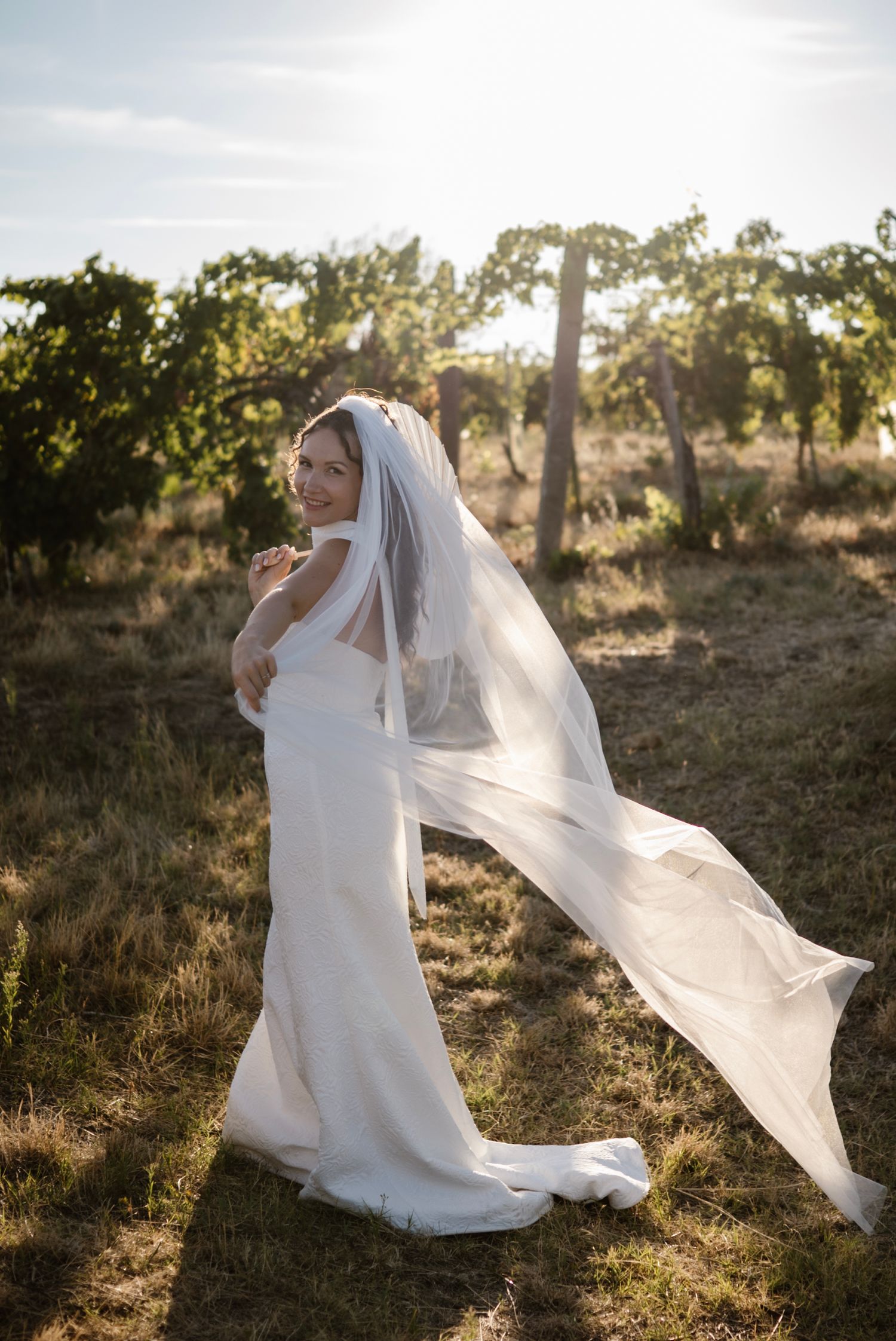 Bride in white gown and flowing veil standing in sunlit vineyard, smiling over shoulder.