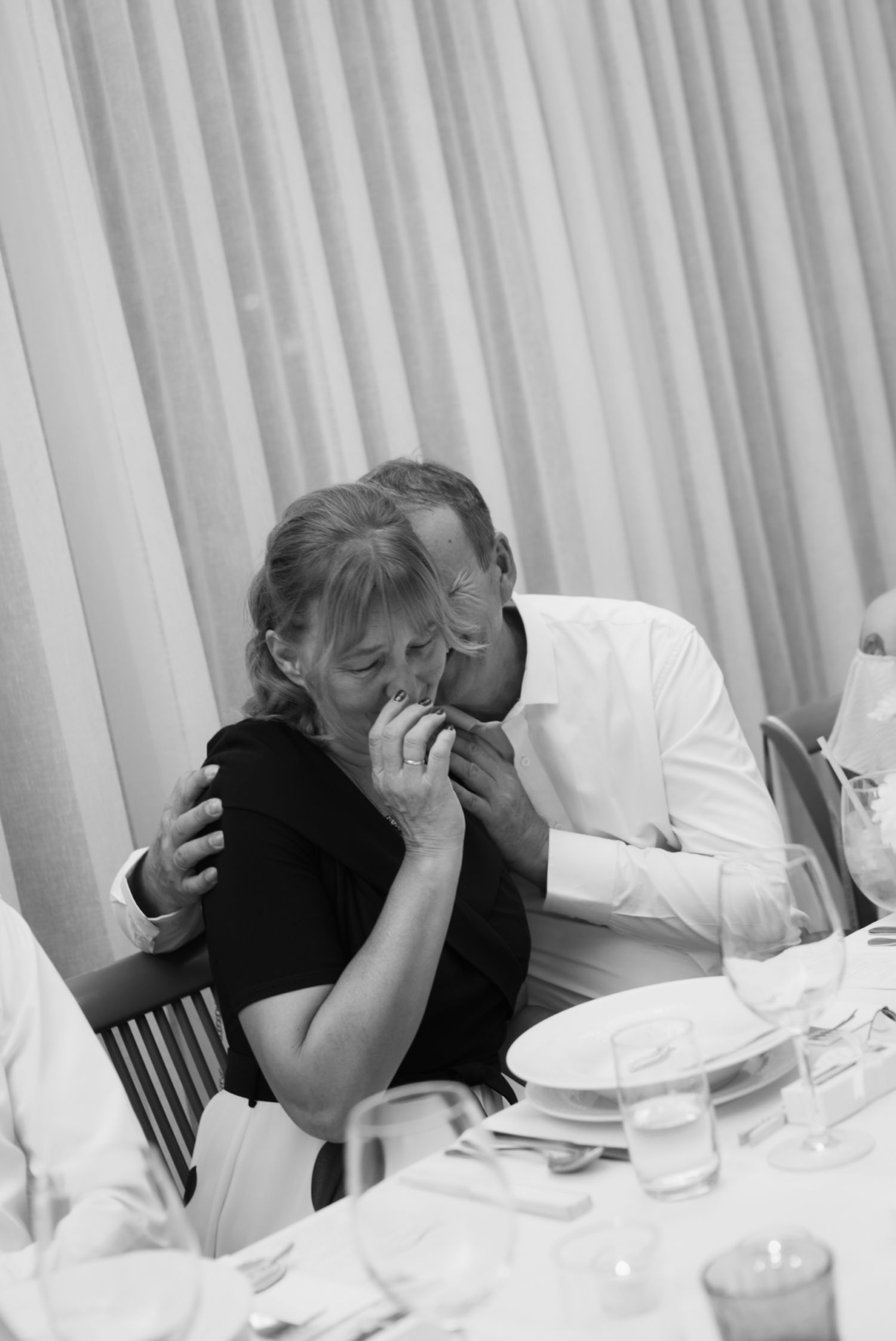 Black and white photo of an emotional couple embracing at a wedding reception dinner table.