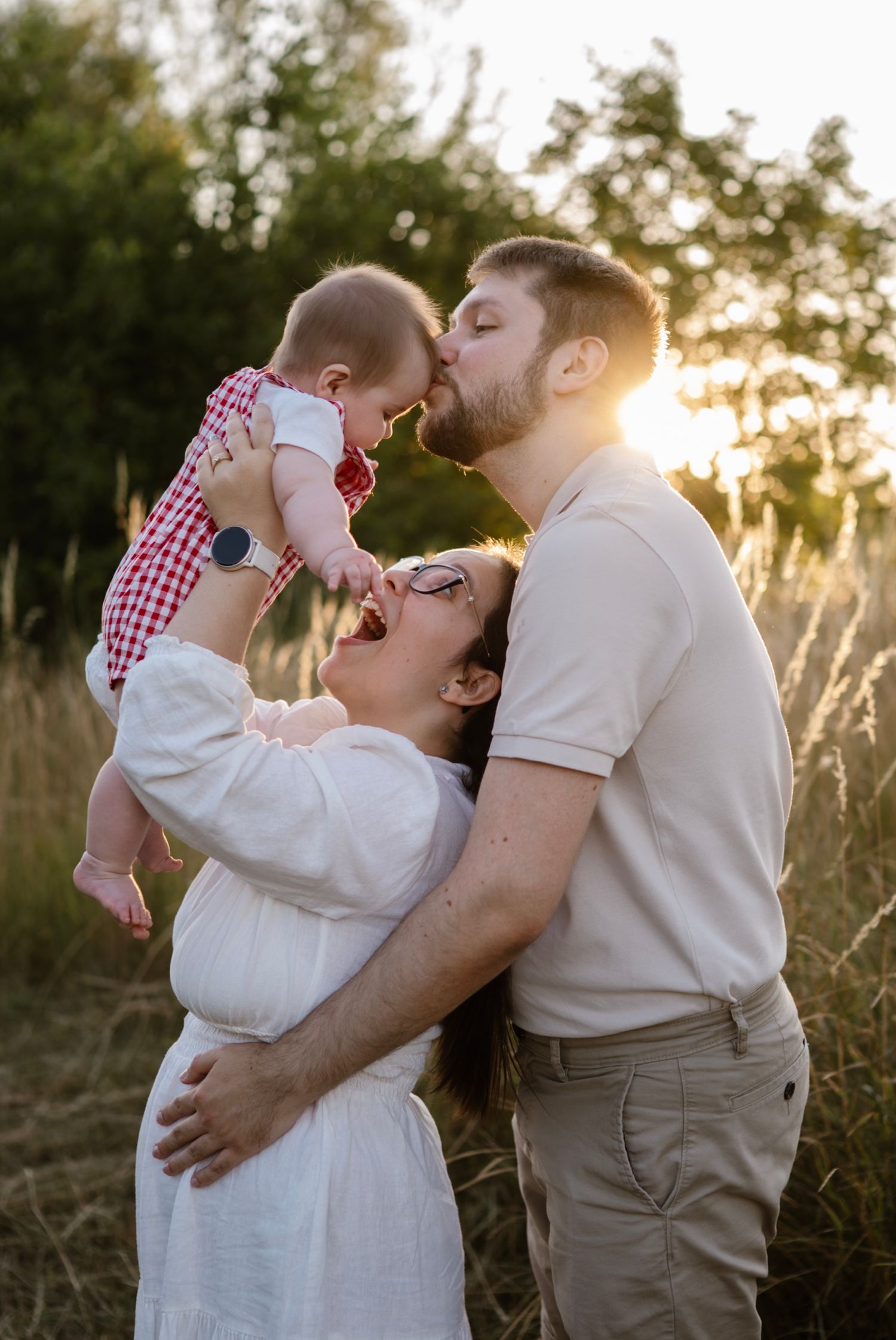 A joyful family poses at golden hour, with parents kissing their baby girl in a sunlit field.