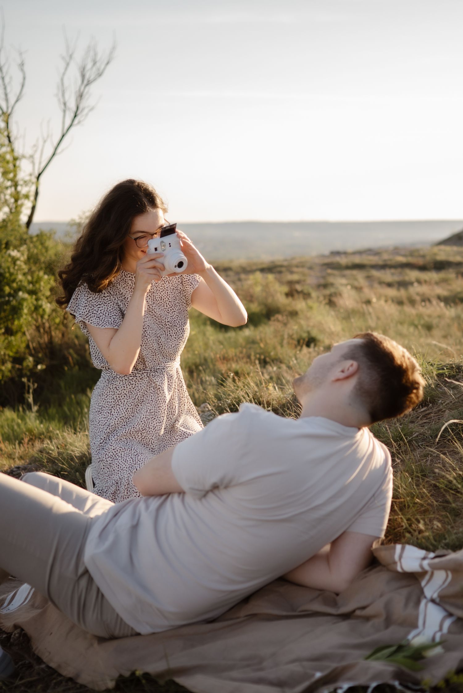 Woman in floral dress photographs man reclining on blanket during golden hour picnic in scenic countryside.