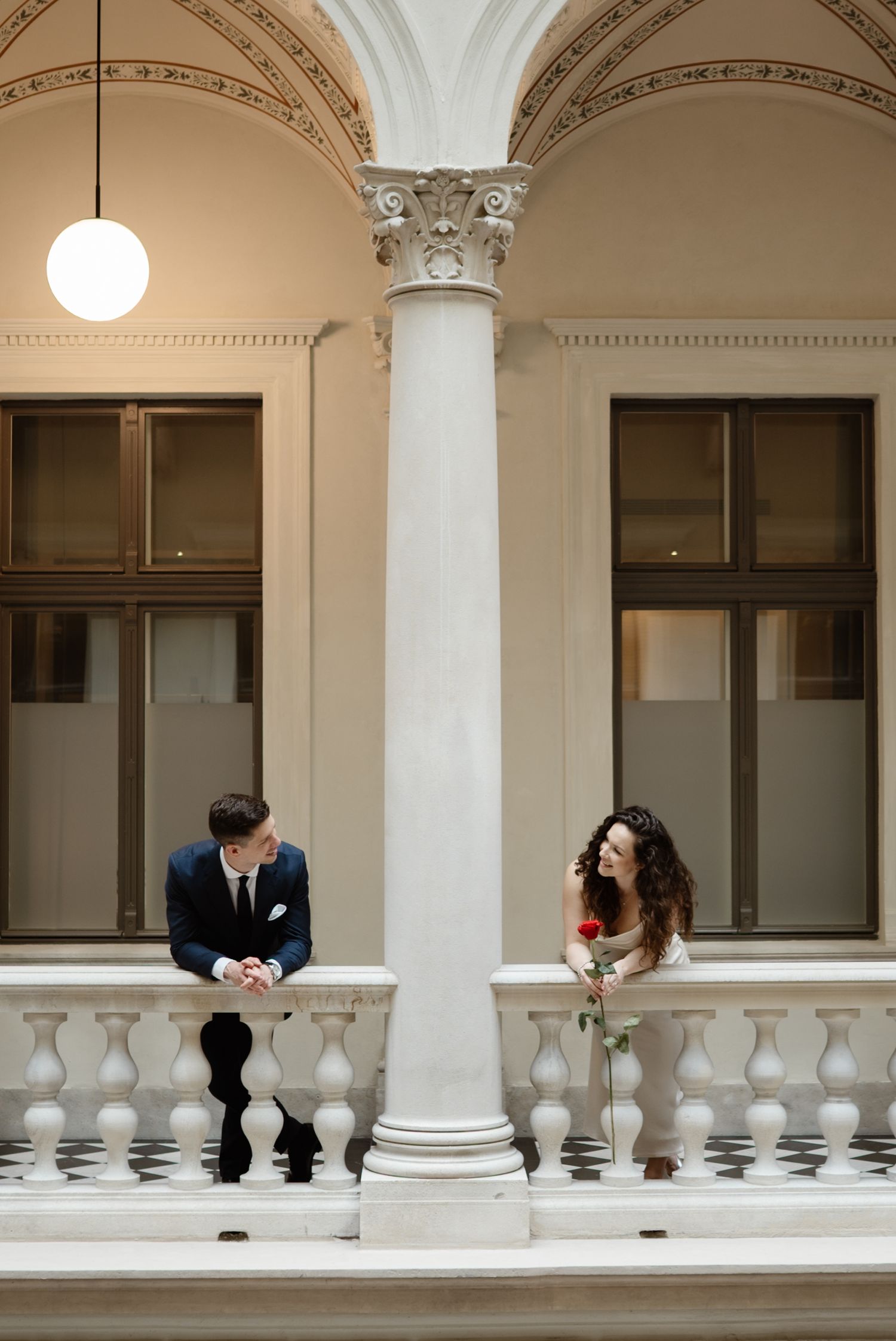 Bride and groom stand on elegant white balcony with ornate columns, sharing a romantic moment with a red rose.