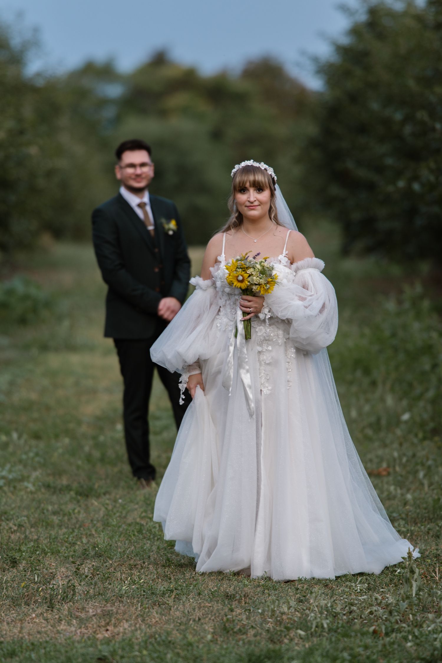 A couple in wedding attire poses in a grassy field with sunflowers.