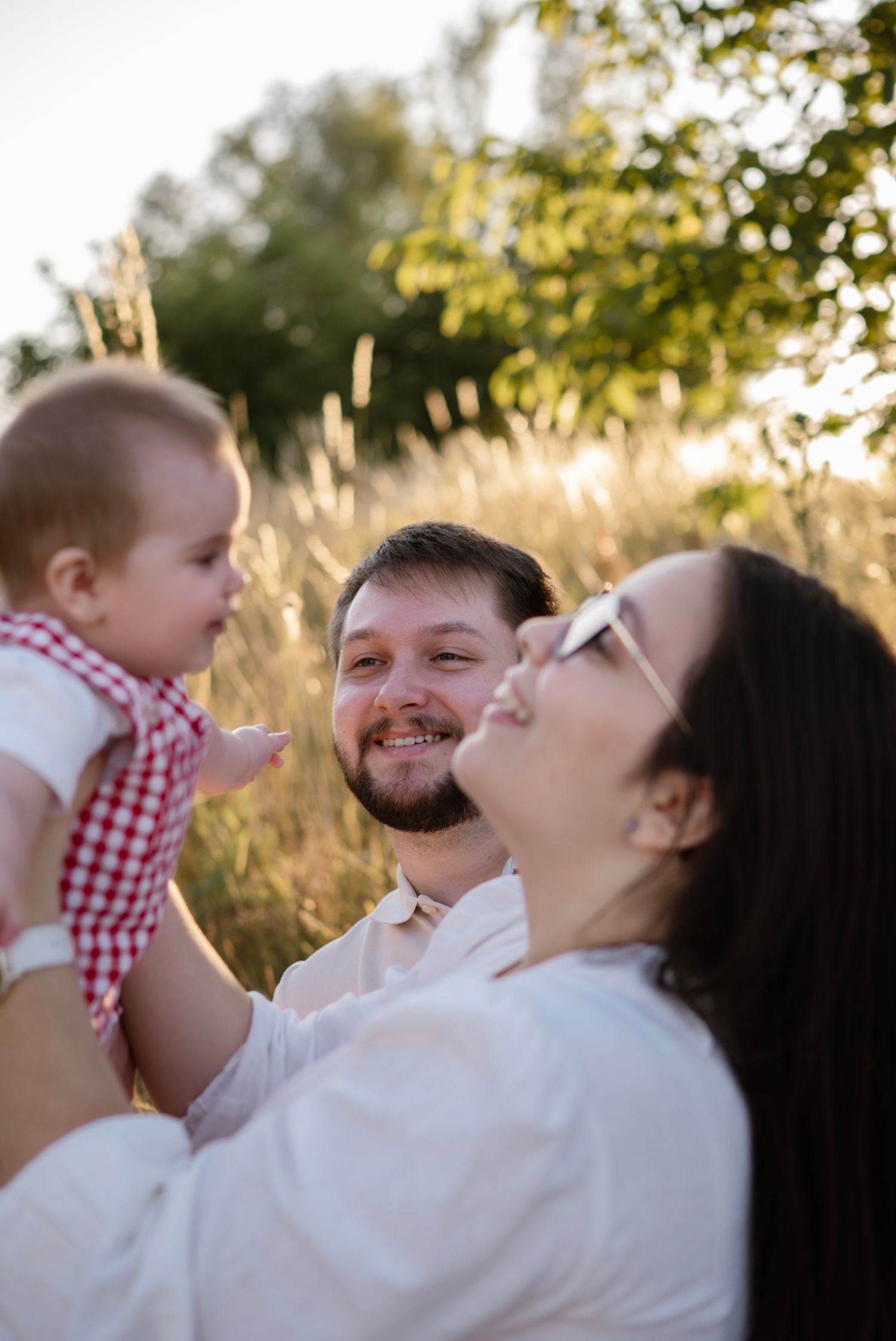 Happy young family with baby outdoors in golden field, parents smiling at infant in red checkered outfit.