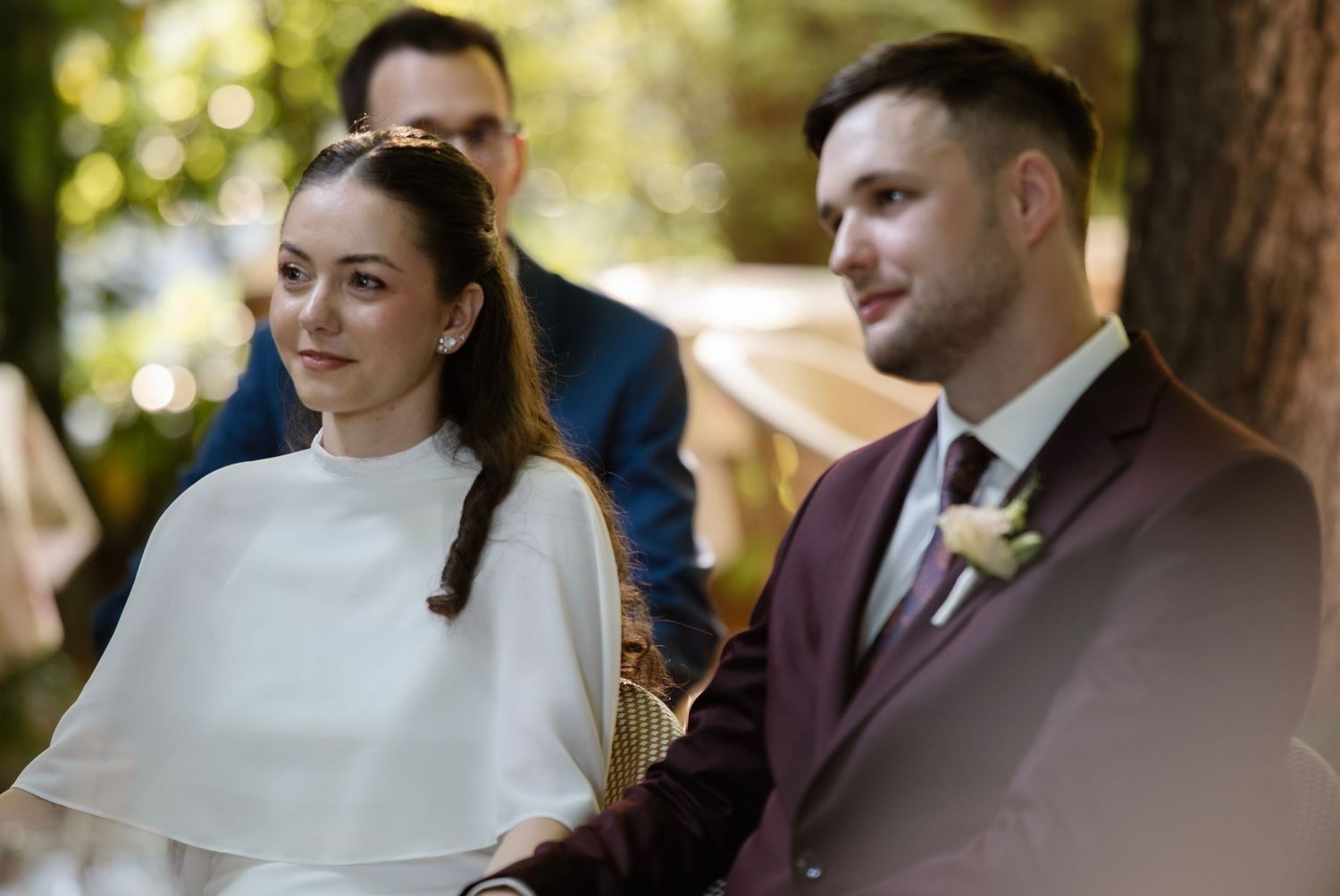 A bride and groom stand together during an outdoor wedding ceremony, smiling softly amid a lush green forest backdrop.