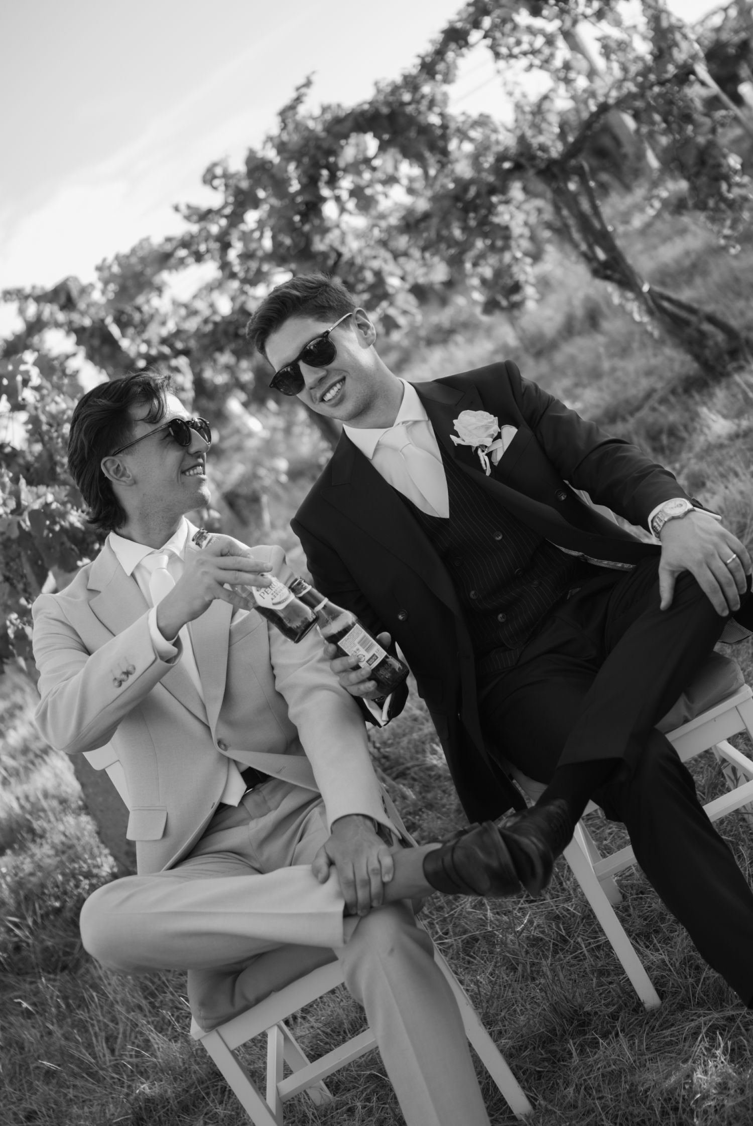 Two grooms in suits and sunglasses share a joyful moment seated outdoors at their wedding ceremony.