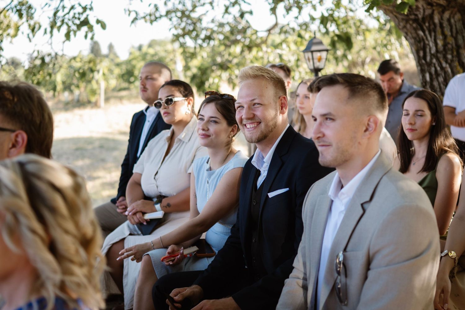 Guests smiling and laughing during an outdoor wedding ceremony seated under trees on a sunny day.