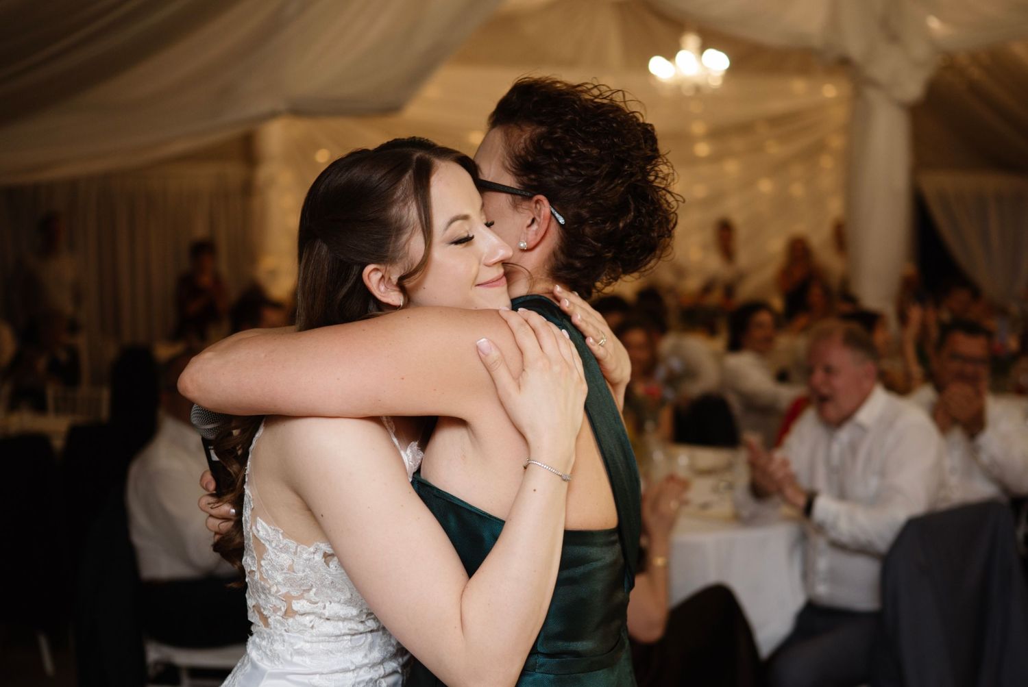 Bride and bridesmaid share an emotional hug during wedding reception in elegant draped tent venue.