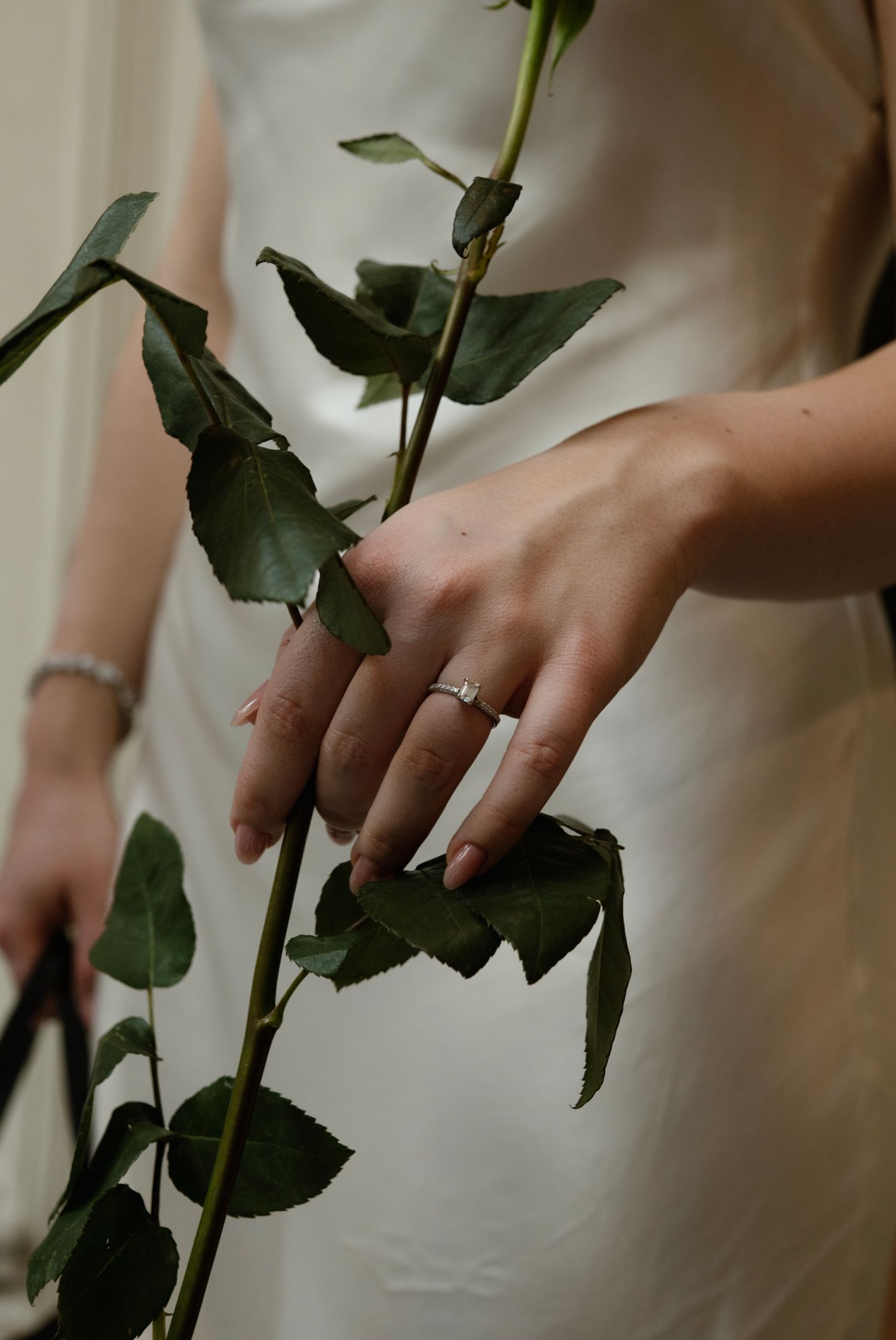 Bride in white dress holding a green rose stem, showcasing a delicate diamond engagement ring on her finger.