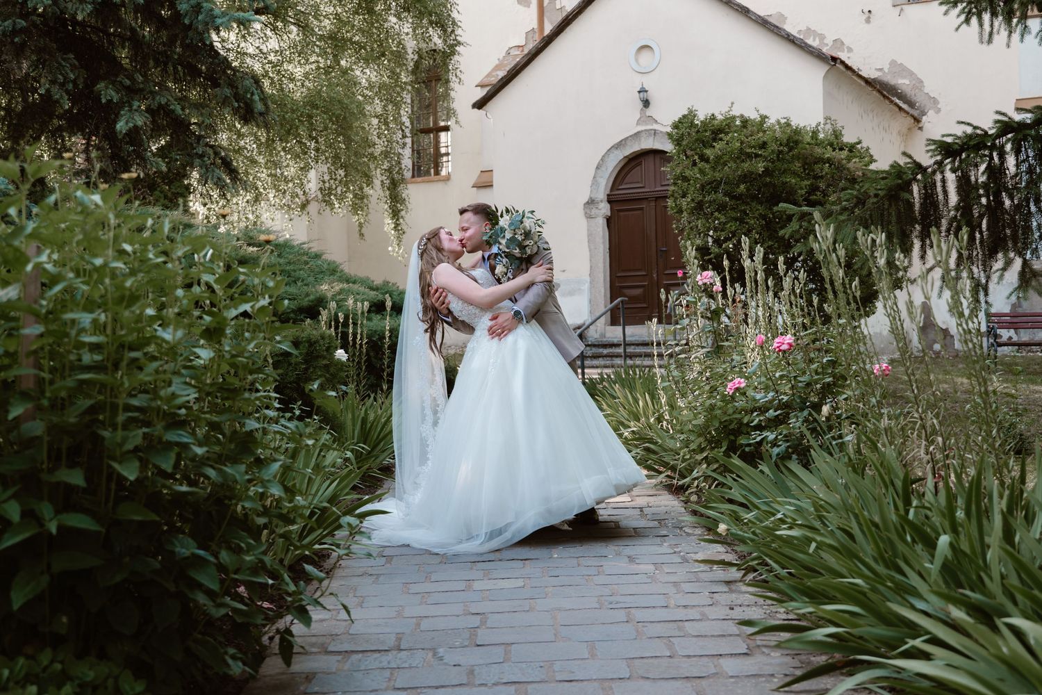 Wedding couple poses in a lush garden pathway outside a white church building.