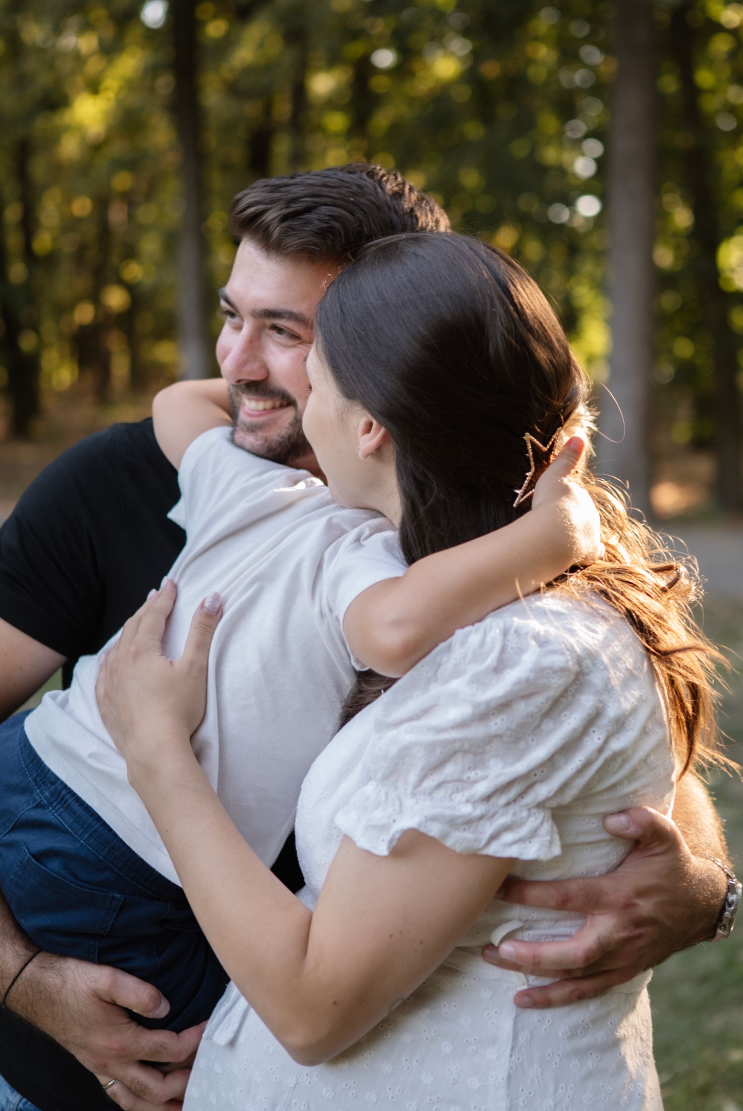 A smiling couple embraces warmly outdoors in a sunlit forest, sharing a joyful moment together.