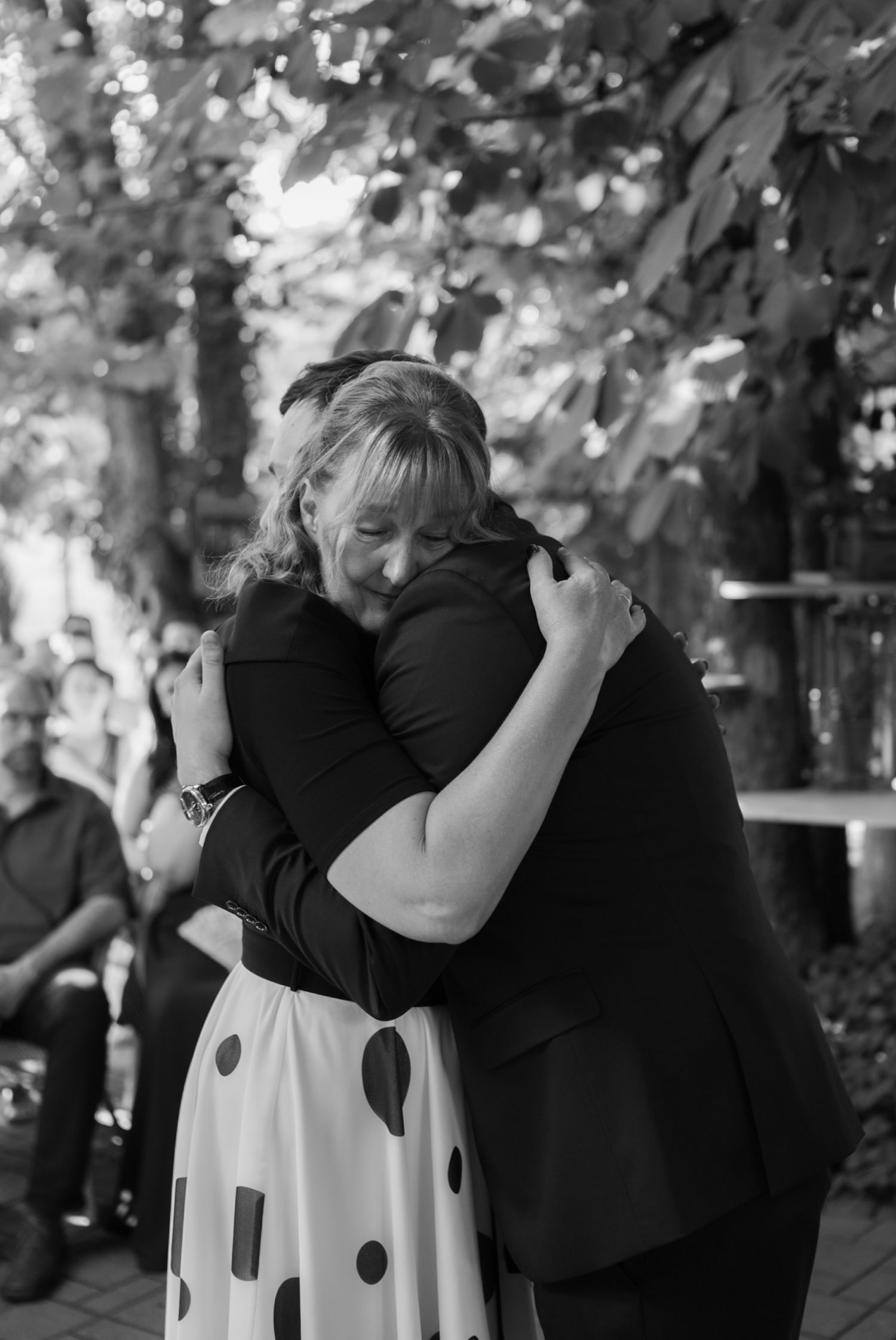 Emotional black and white photo of two wedding guests sharing a heartfelt hug outdoors under leafy trees.