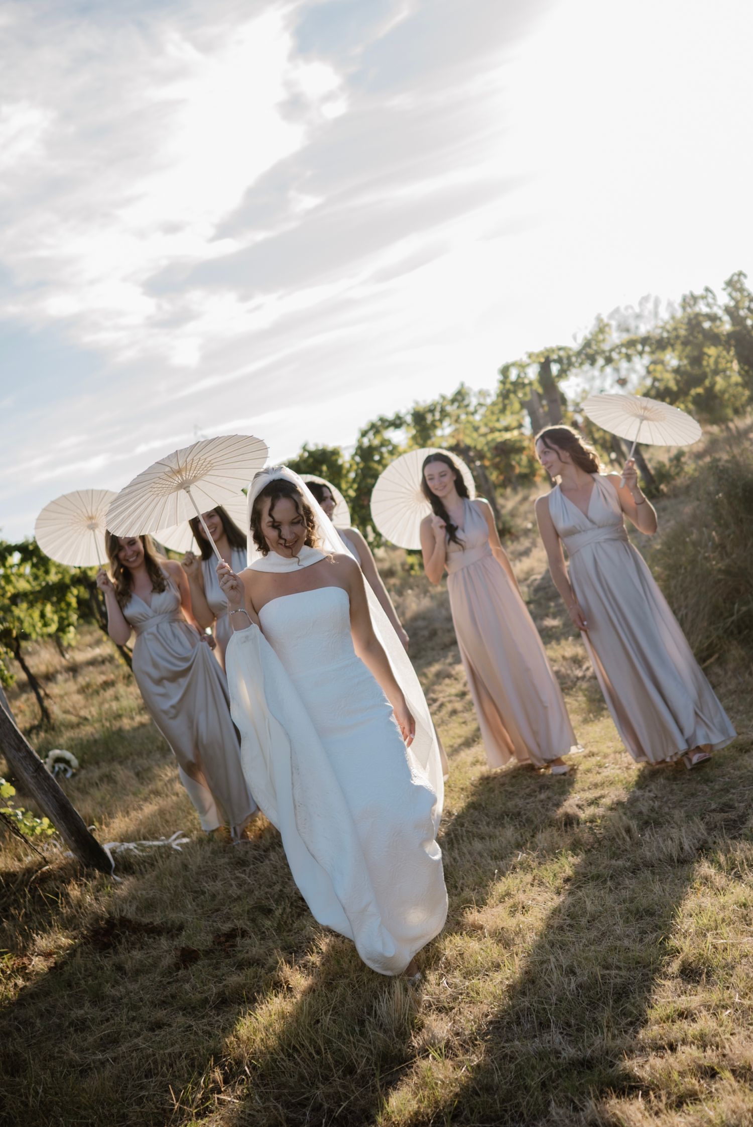 Bride in white gown leads bridesmaids in blush dresses holding parasols through a sunlit vineyard.