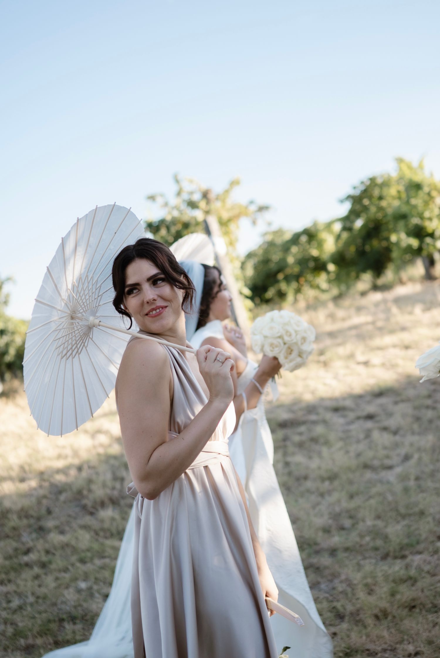 Bridesmaid in champagne dress holds white parasol and bouquet at outdoor wedding ceremony.