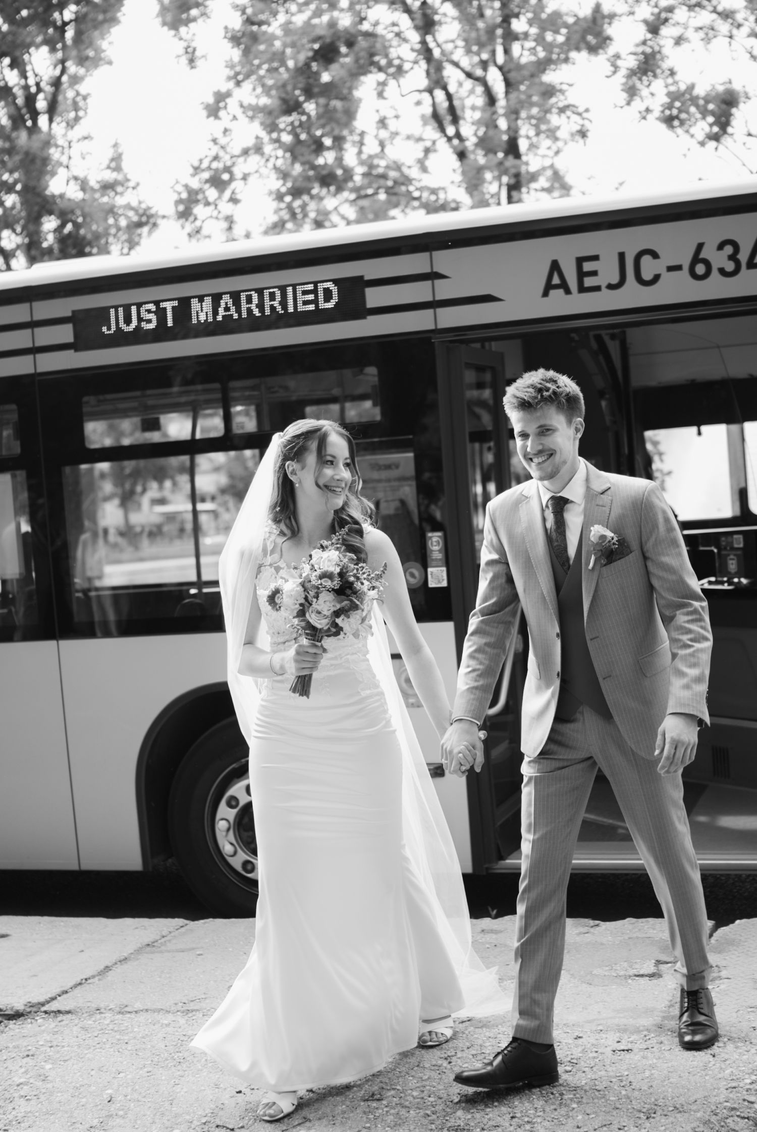 Just married couple holding hands and smiling in front of a bus labeled Just Married in black and white.