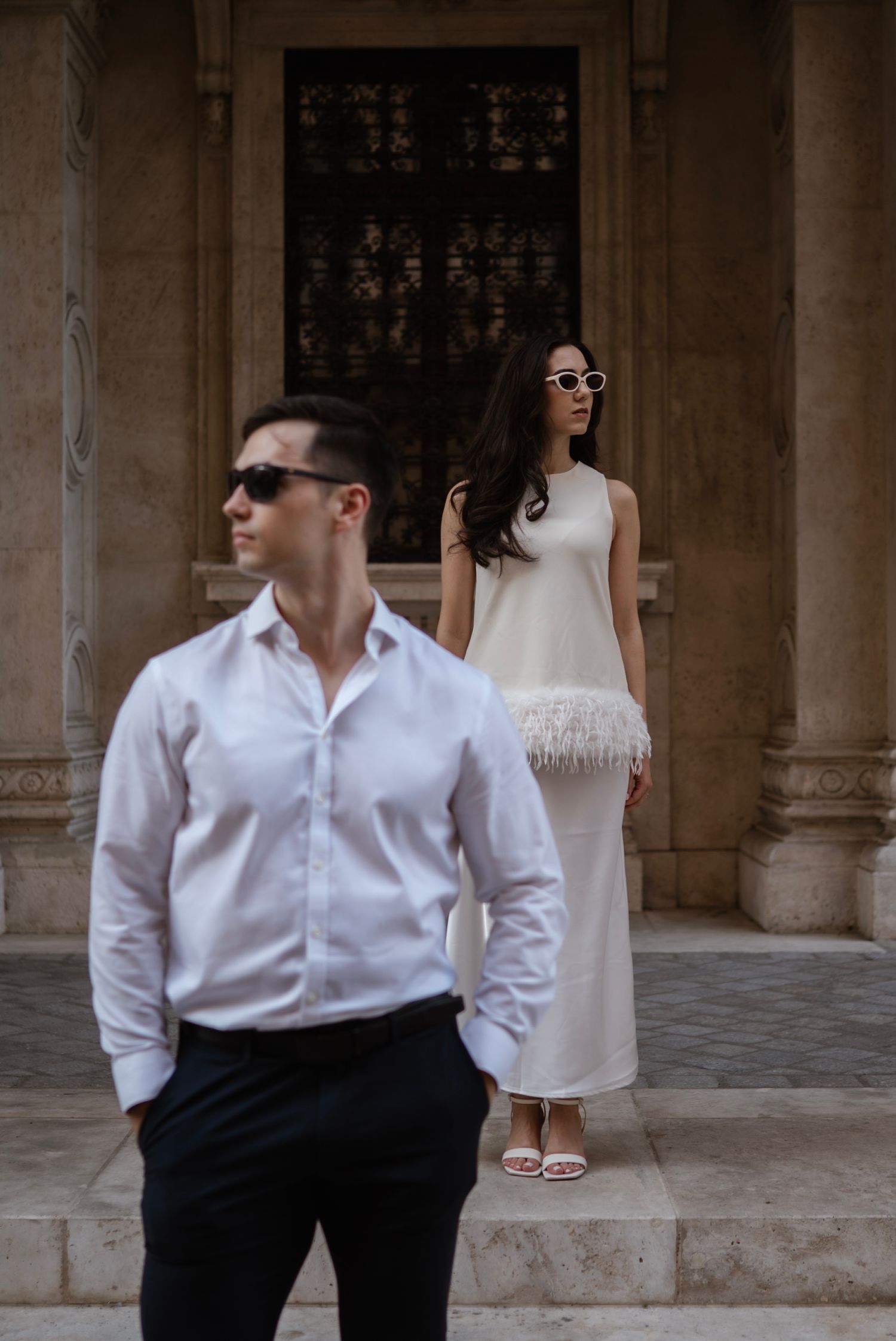 Elegant couple in white outfits posing outside ornate building, woman in feathered dress with sunglasses, man in white shirt.