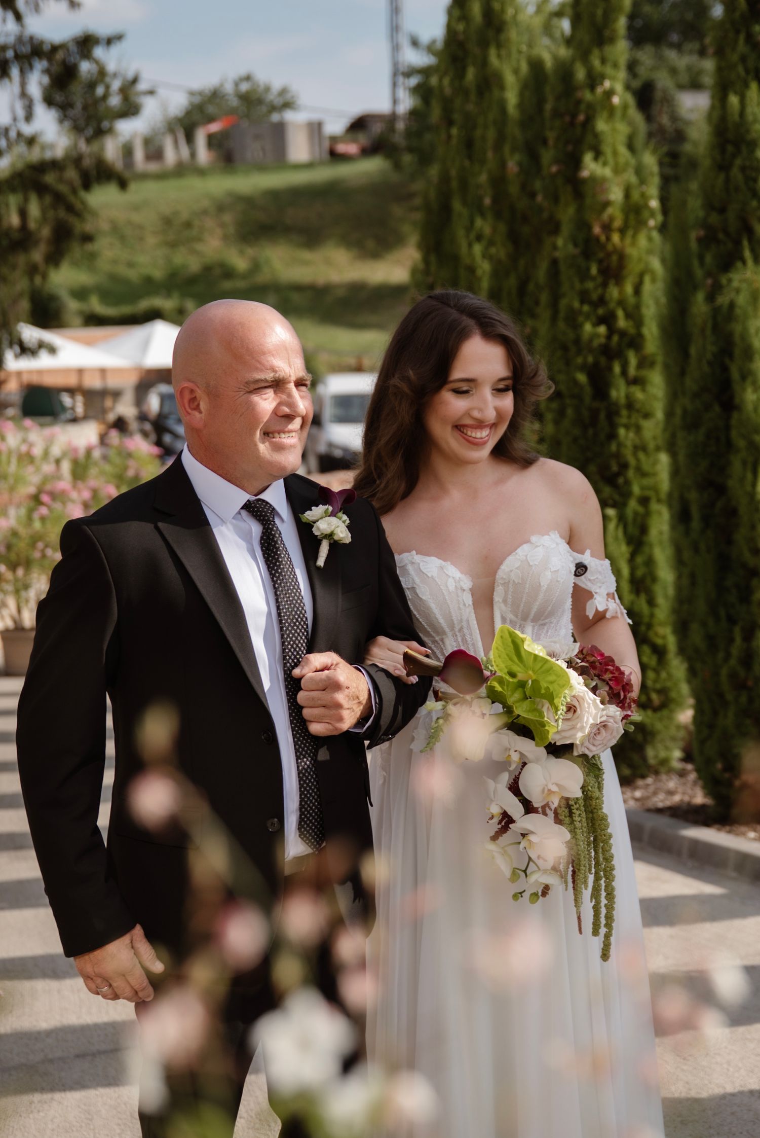 Smiling bride in white off-shoulder gown walks down the aisle with bald escort in black tuxedo.