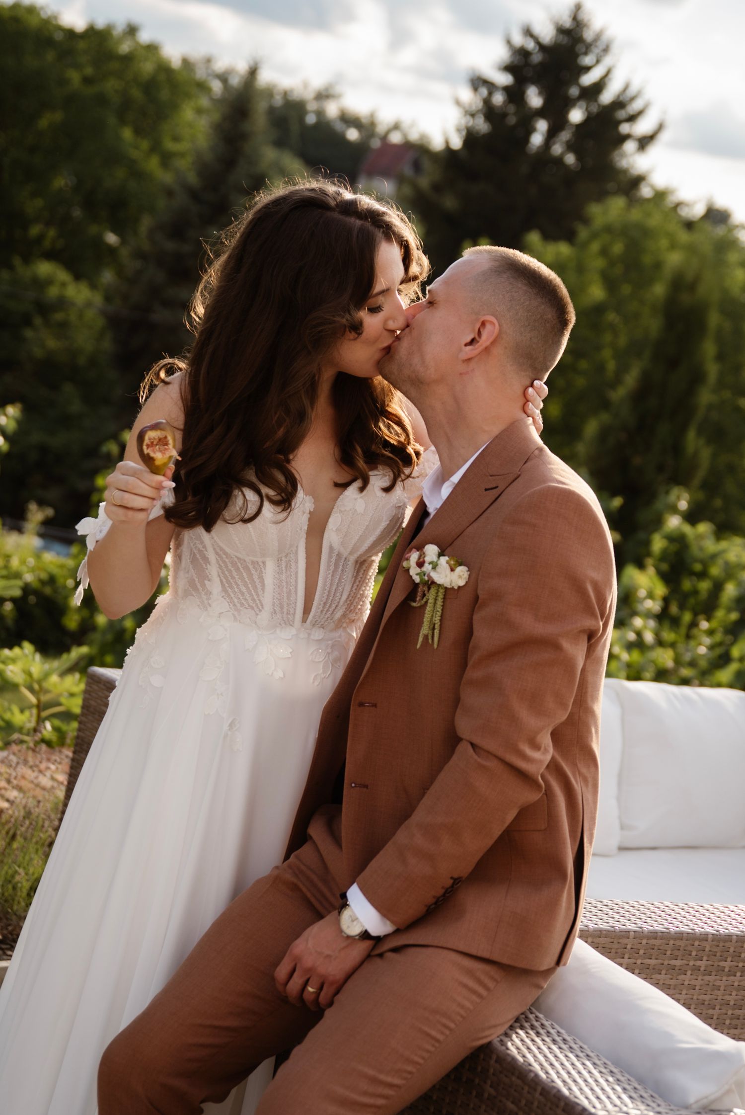 A bride and groom share a romantic kiss outdoors, holding champagne glasses, dressed in white gown and tan suit.