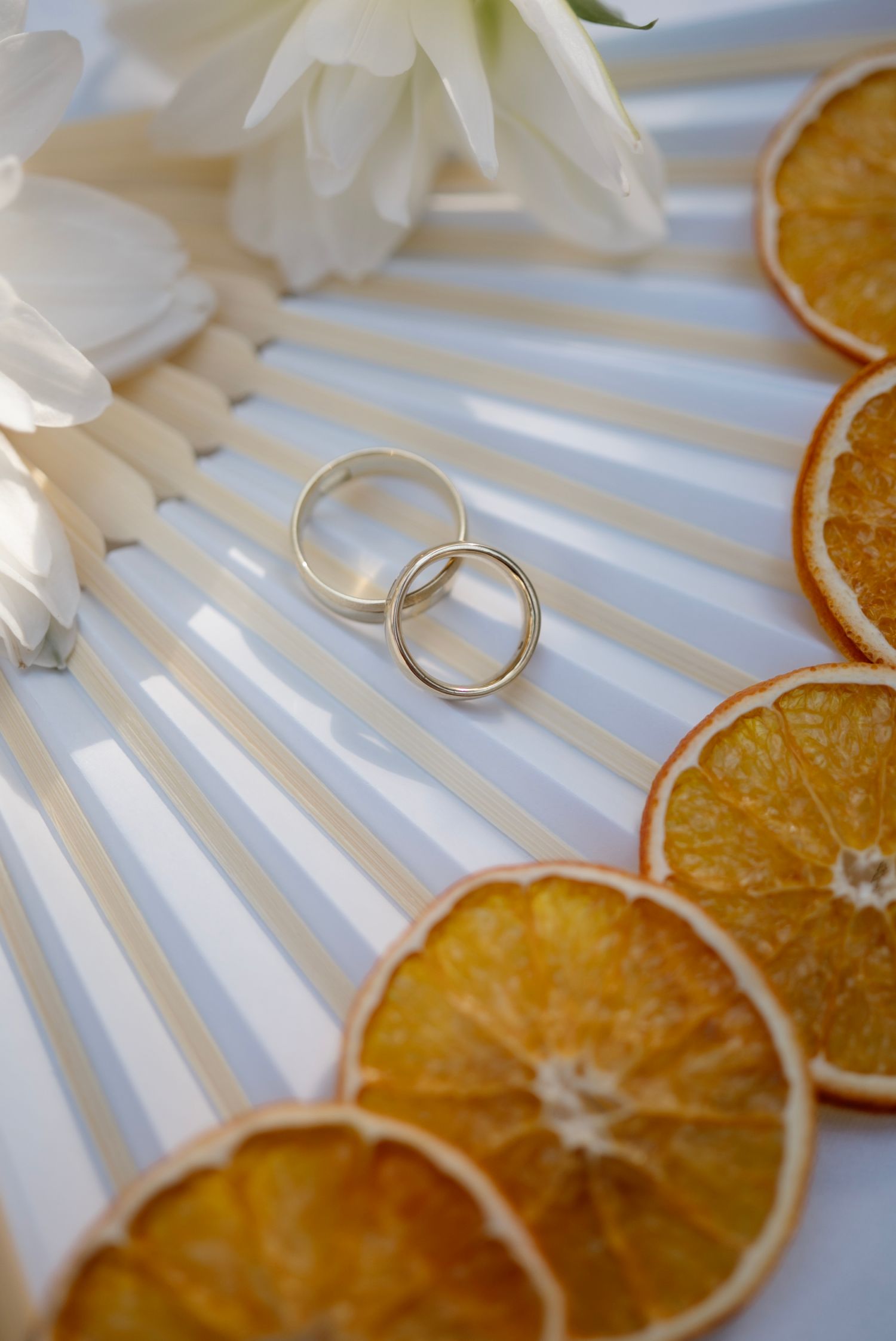 Two gold wedding bands rest on a white fan beside dried orange slices and white flowers.