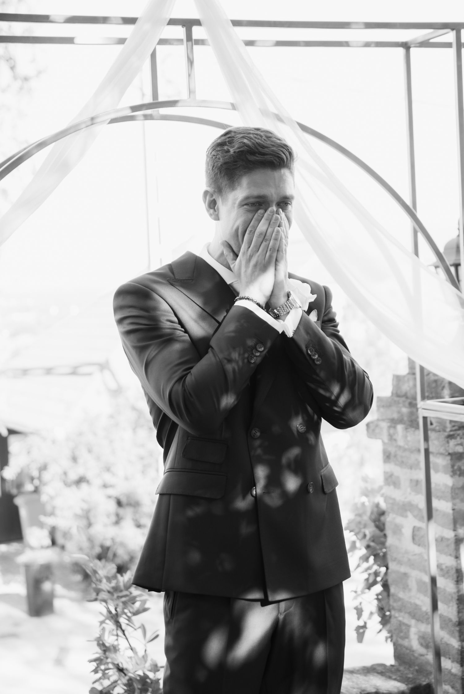 Emotional groom in black suit covering his face with hands at wedding altar in black and white.