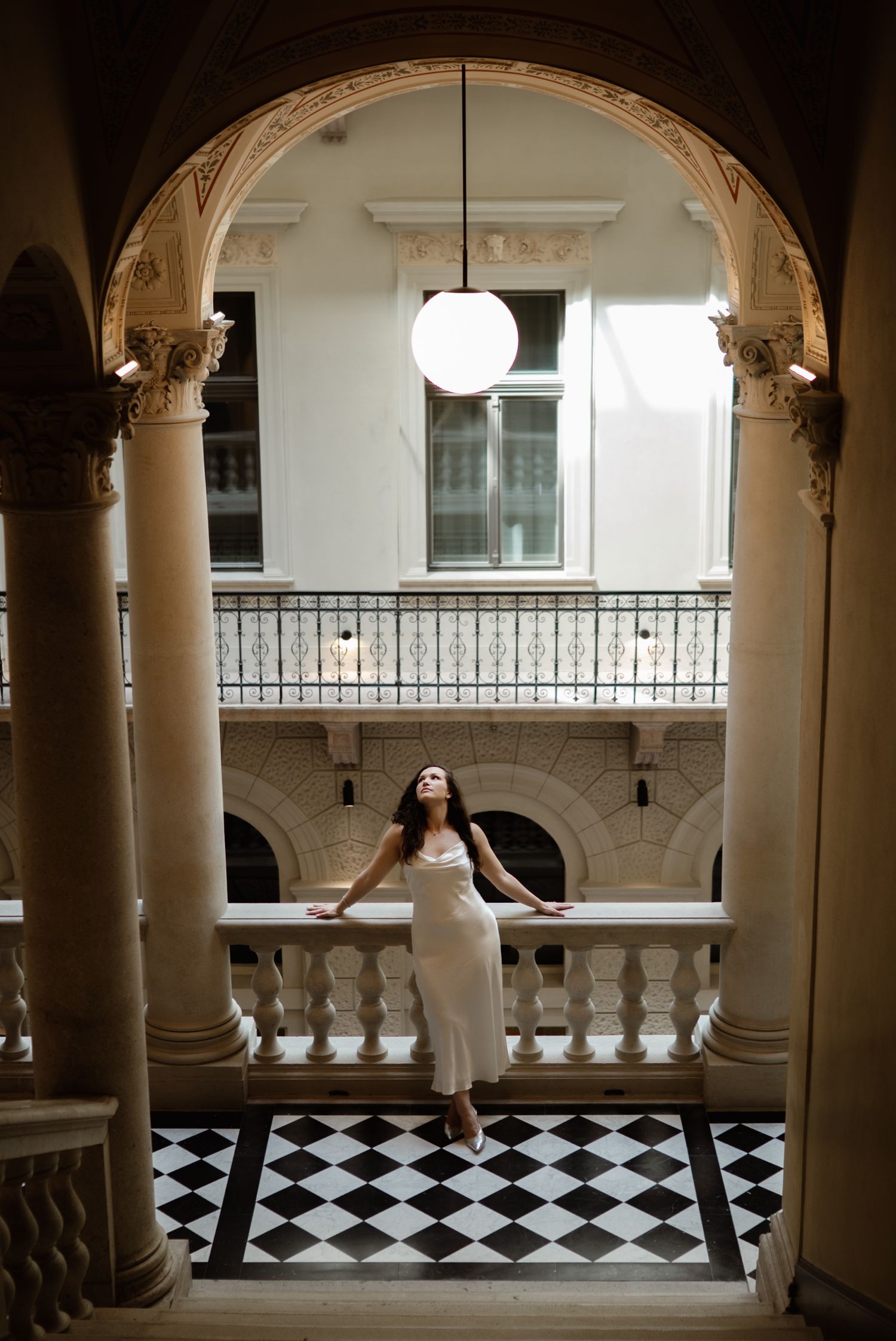 Woman in white dress posing gracefully on a checkered floor beneath an elegant arched colonnade.