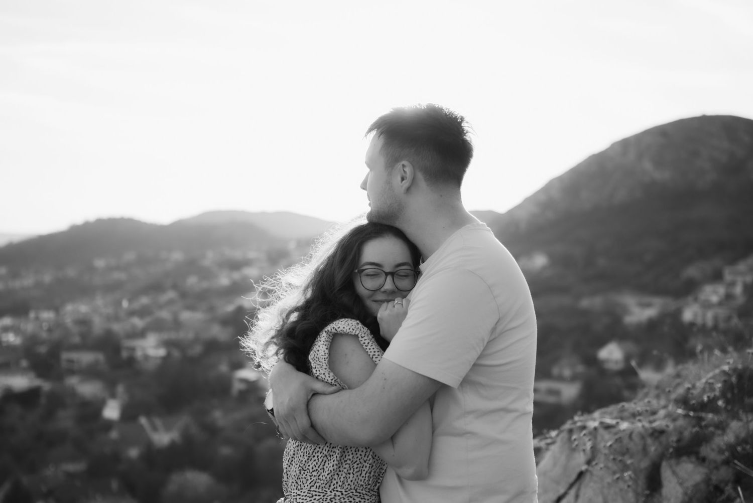 Black and white photo of a couple embracing outdoors with scenic mountain and town views in the background.