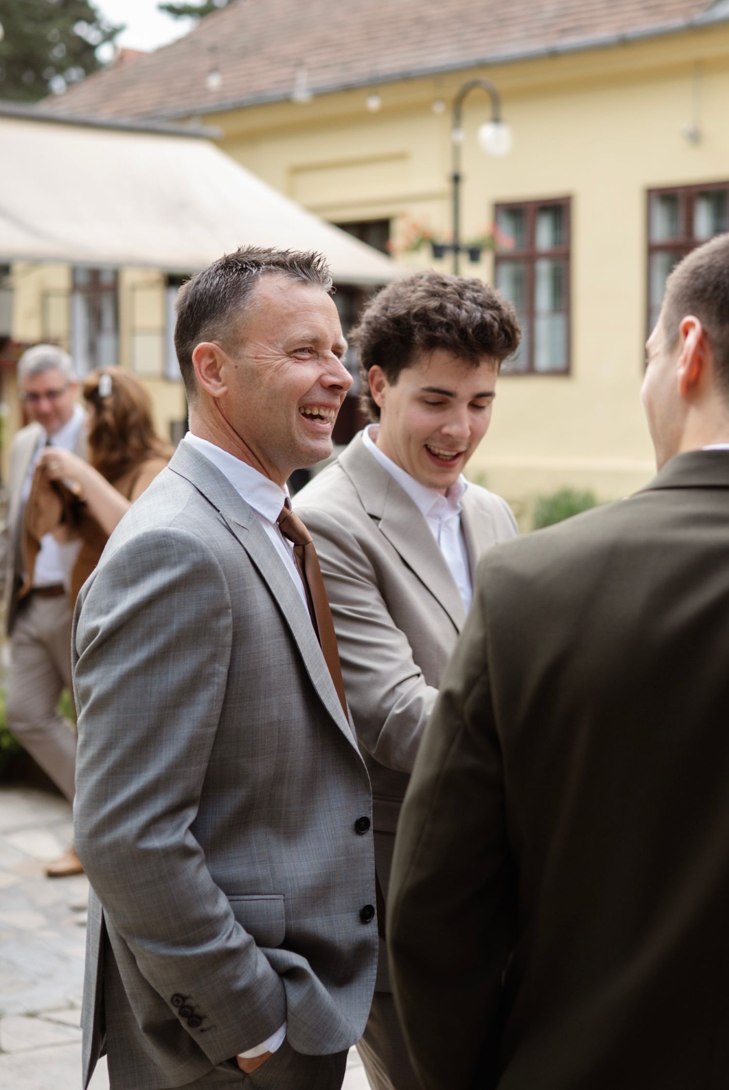 Two men in gray suits laughing and conversing outdoors at a wedding reception near a yellow building.