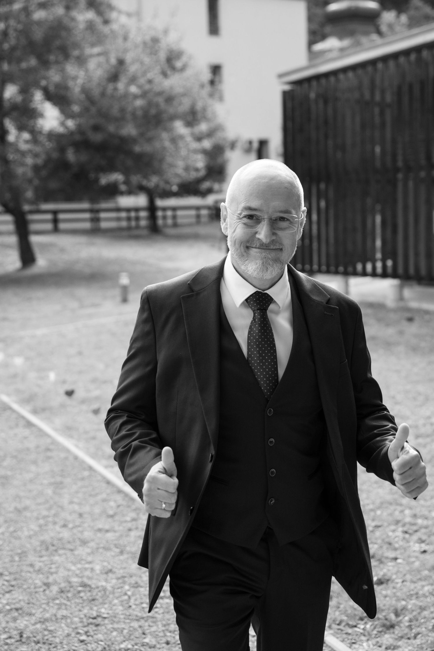 A gentleman in a three-piece suit gives two thumbs up while smiling outdoors.