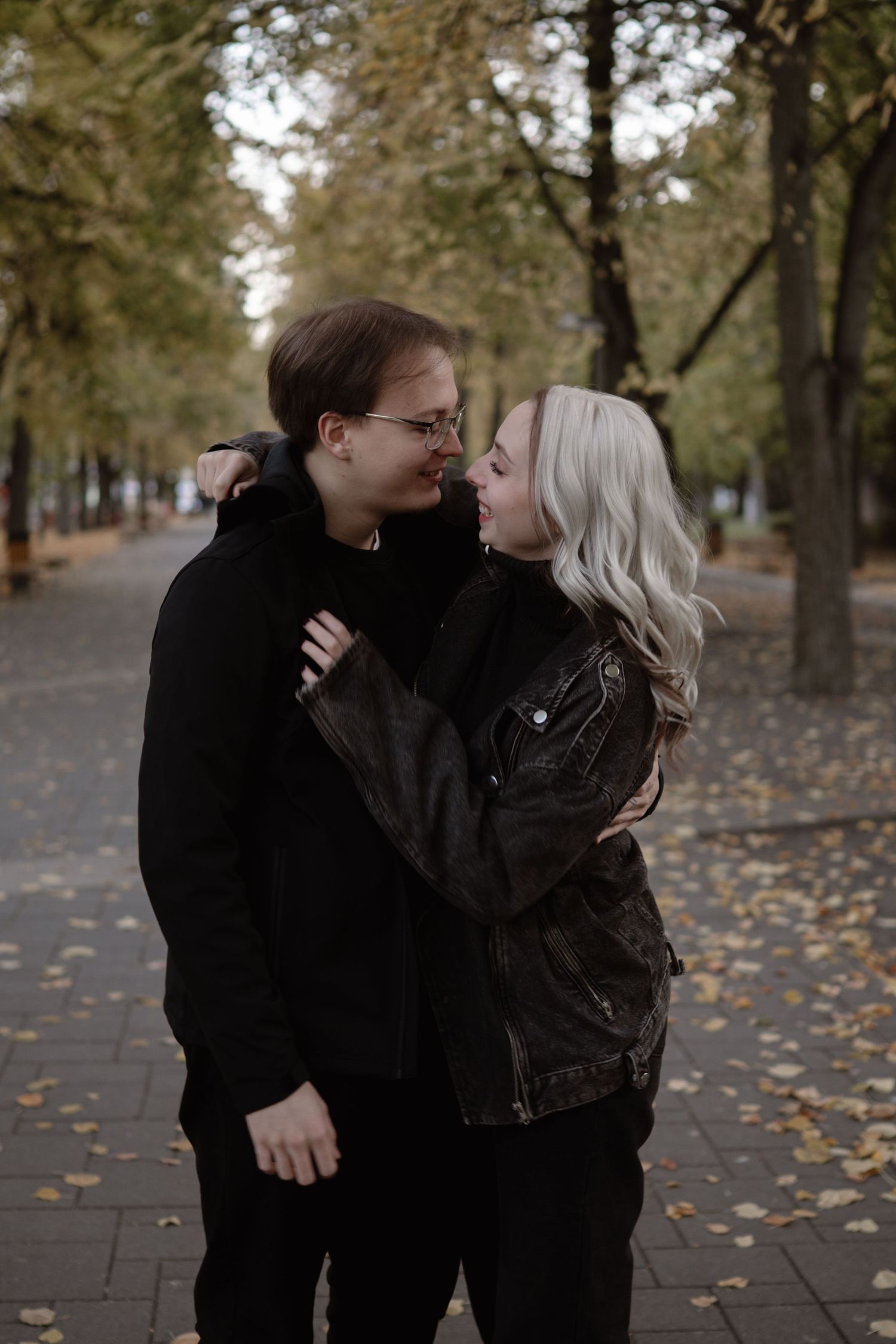 Couple in black jackets embracing on leaf-covered path during fall.