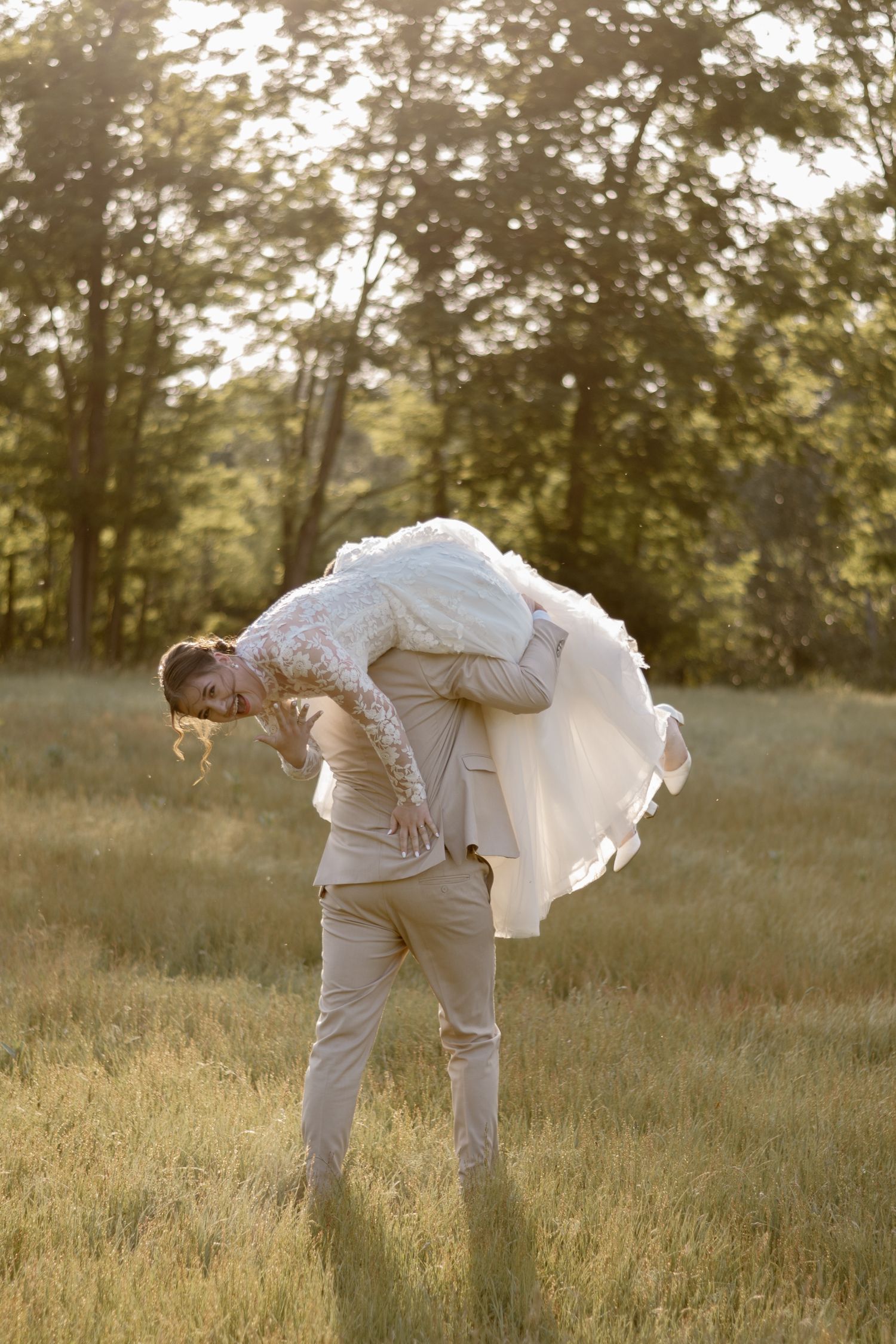 A groom carries his bride playfully in a sunlit meadow at golden hour.