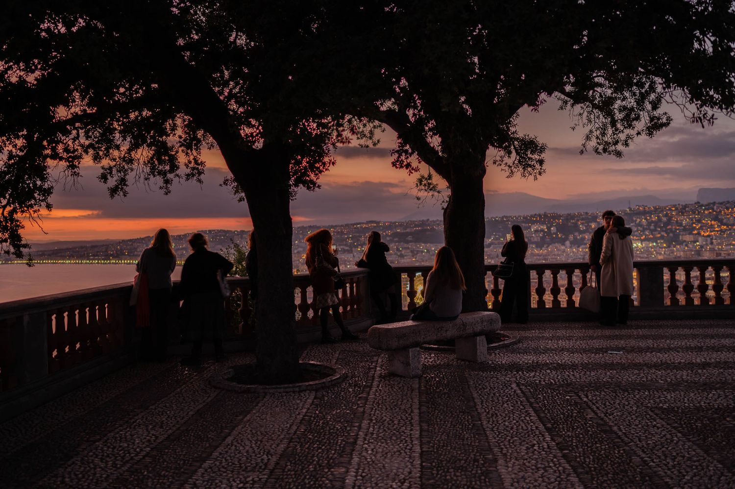 Silhouettes of people gathered on a terrace overlooking a sunset view through trees.