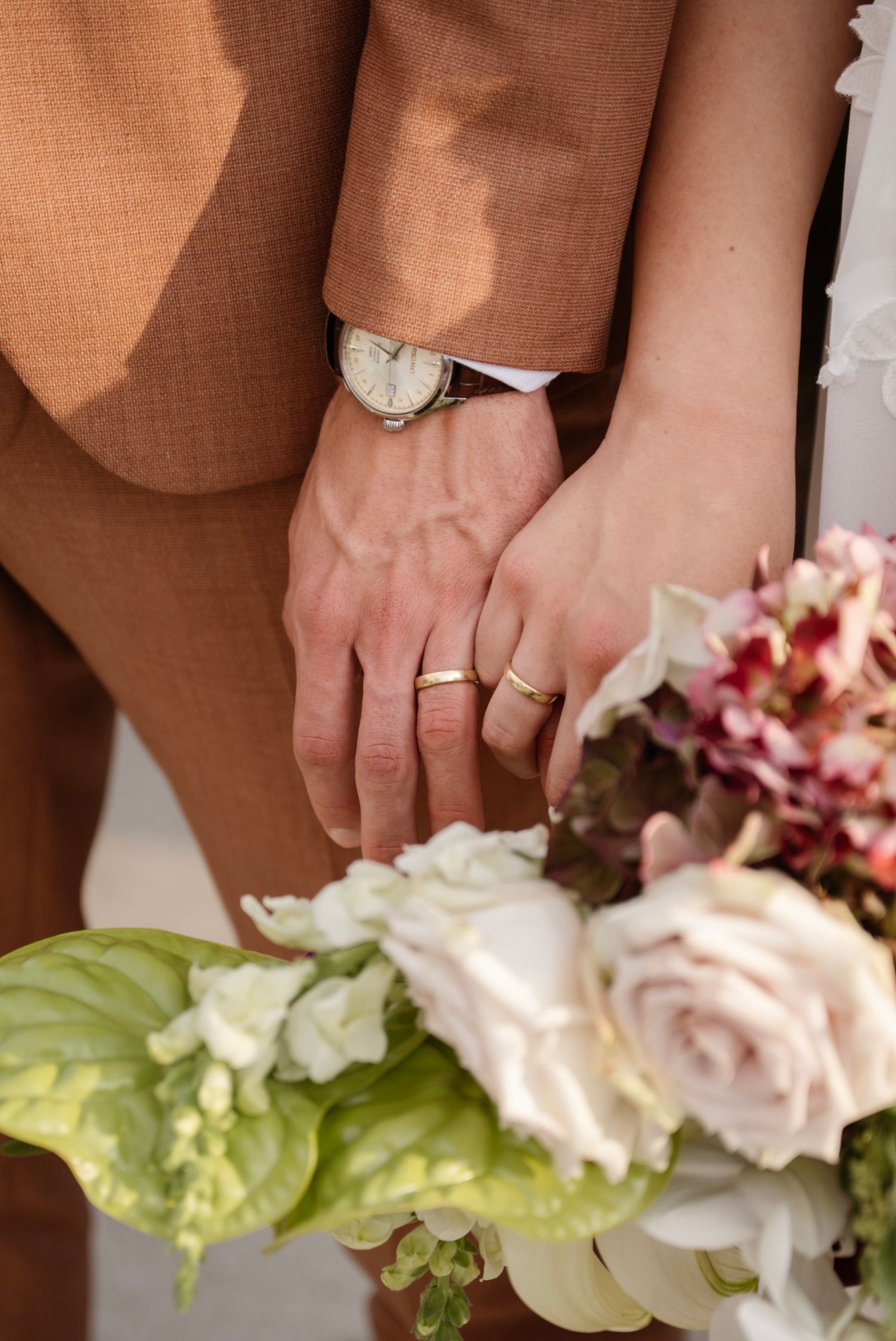Newlywed couple holding hands showing gold wedding rings, with a bridal bouquet of pink roses and white flowers.